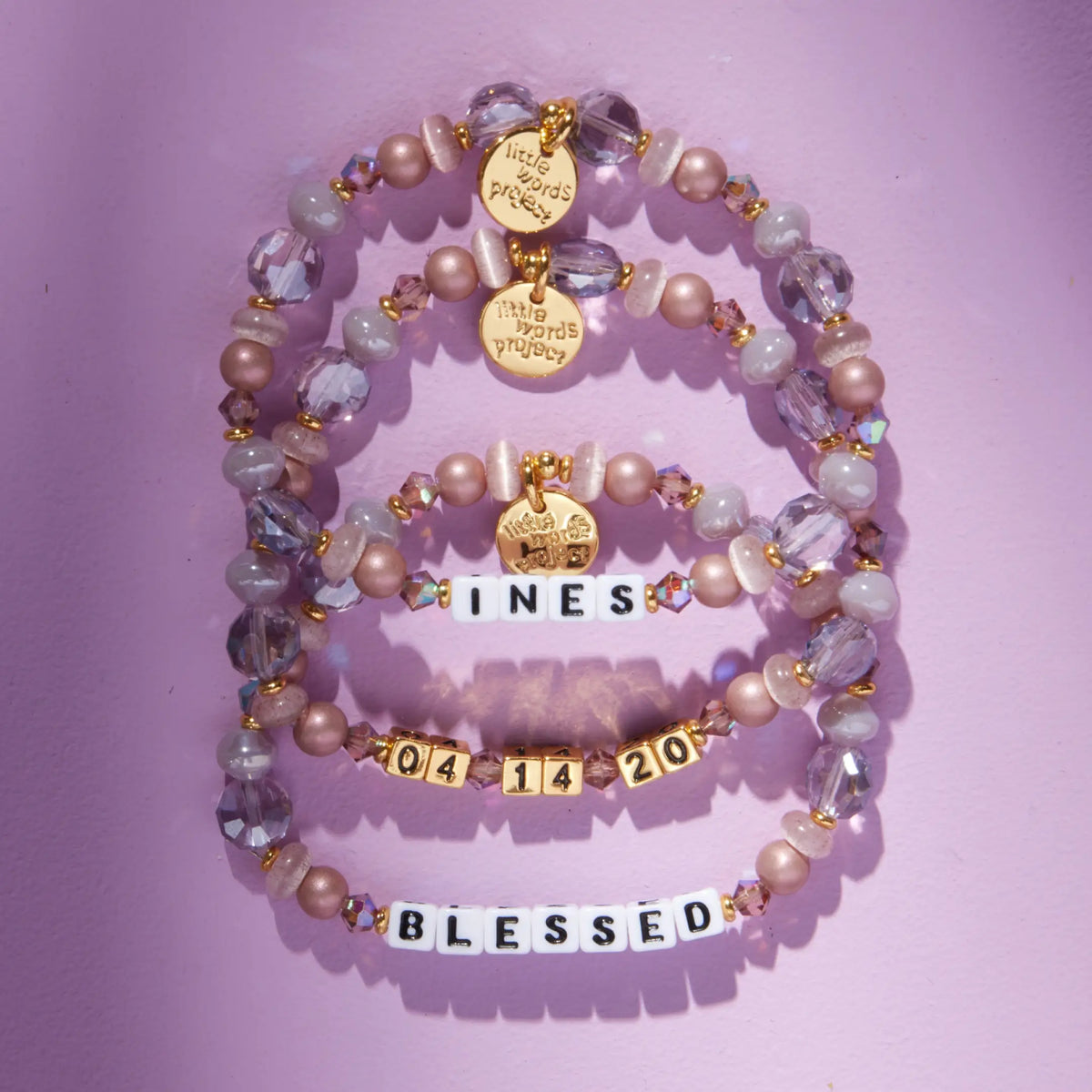 Three Custom Nice Spice bracelets reading Ines, 04 14 20, and Blessed in white and gold letter beads, mauve pearl and lavender crystal beads, flat-lay on lavender background