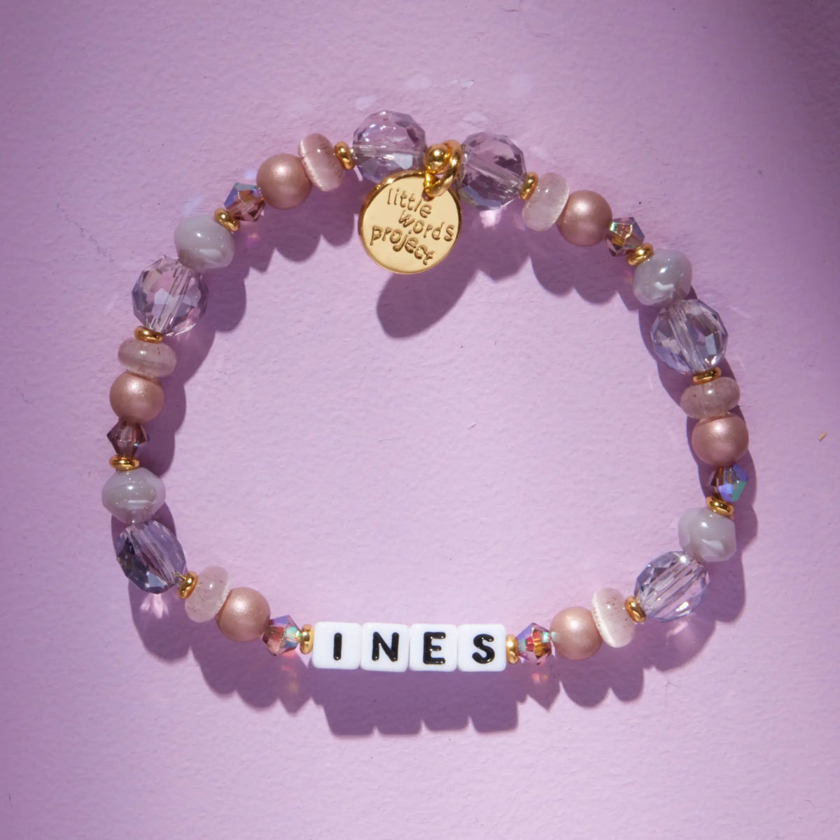 Custom Nice Spice bracelet with white letter beads reading Ines, mauve pearl, lavender crystal, and clear faceted beads, flat-lay on lavender background