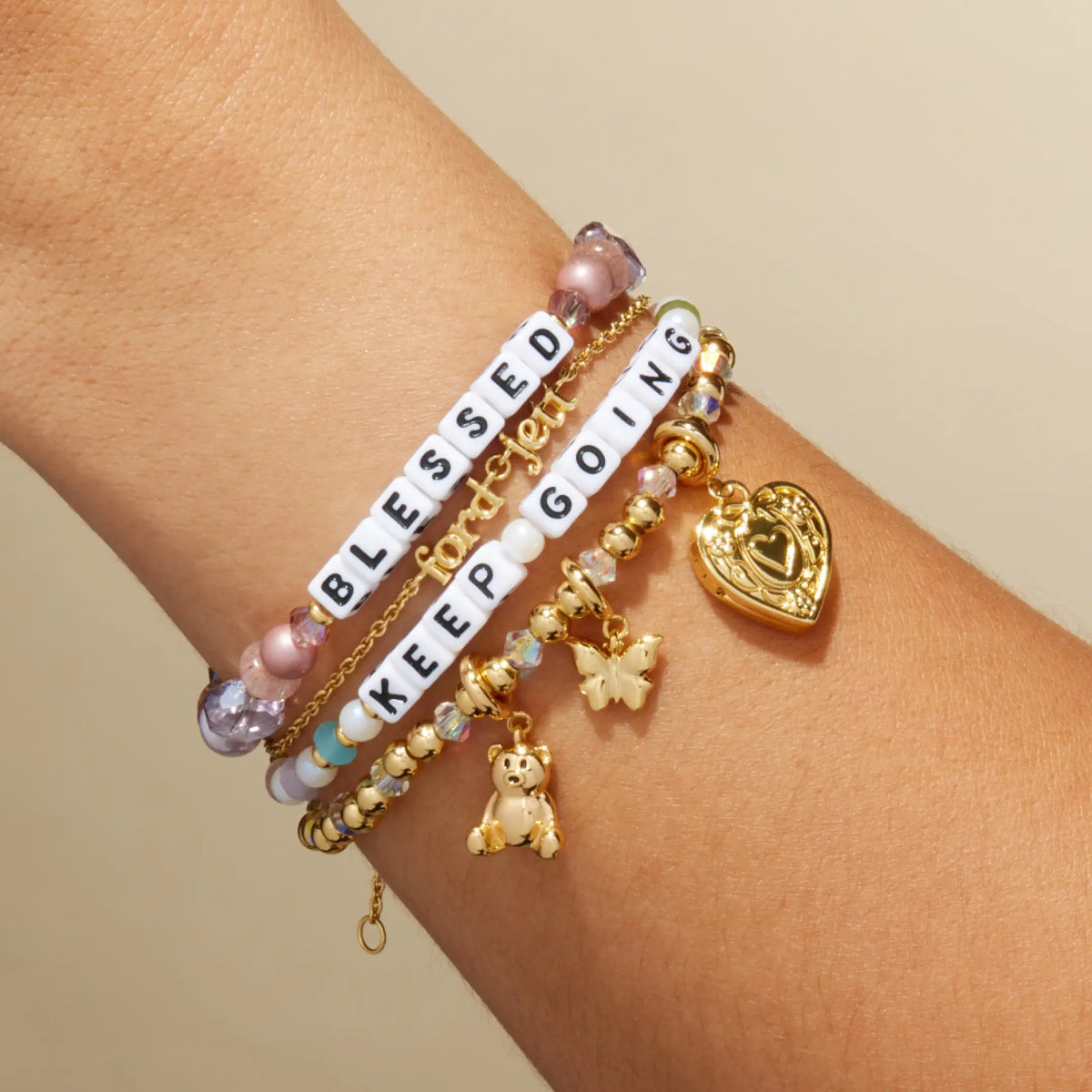Custom Nice Spice bracelet with white letter beads reading Blessed stacked with a Keep Going bracelet and gold charm bracelets, on-model on wrist against a tan background