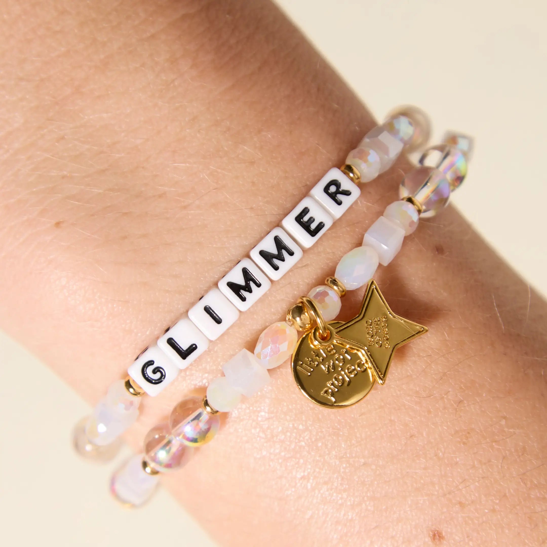 Bracelet with 'GLIMMER' charm on wrist against beige background