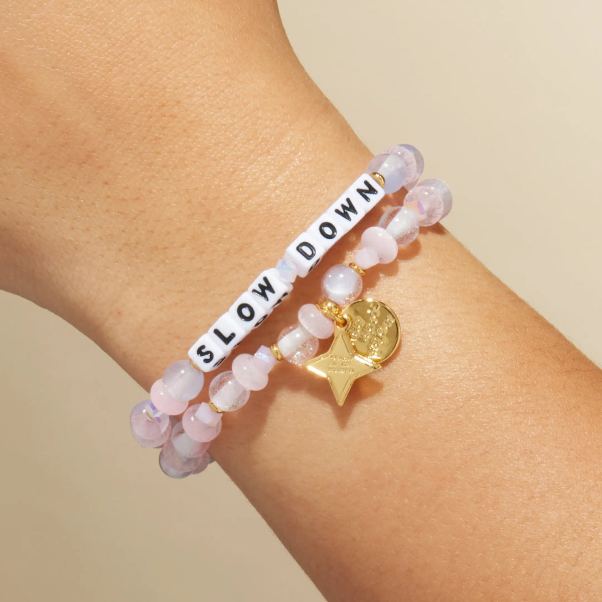 Slow Down October Word of the Month bracelet with white letter beads, pale pink glitter glass and cat eye beads, and a gold star Word of the Month charm, stacked on-model on wrist against cream background