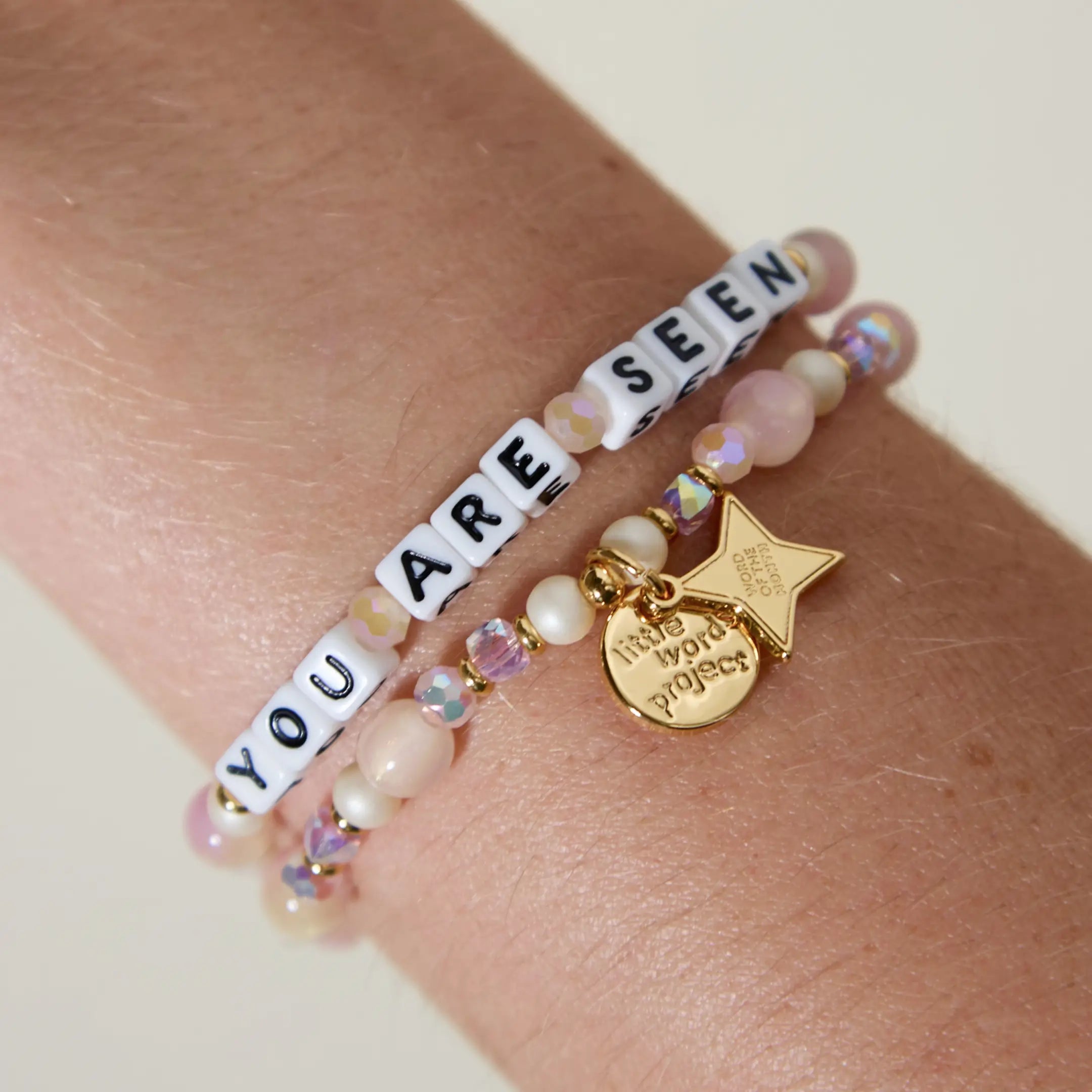 You Are Seen Word of the Month bracelets with white letter beads stacked on a wrist with gold Little Words Project disc and star charms