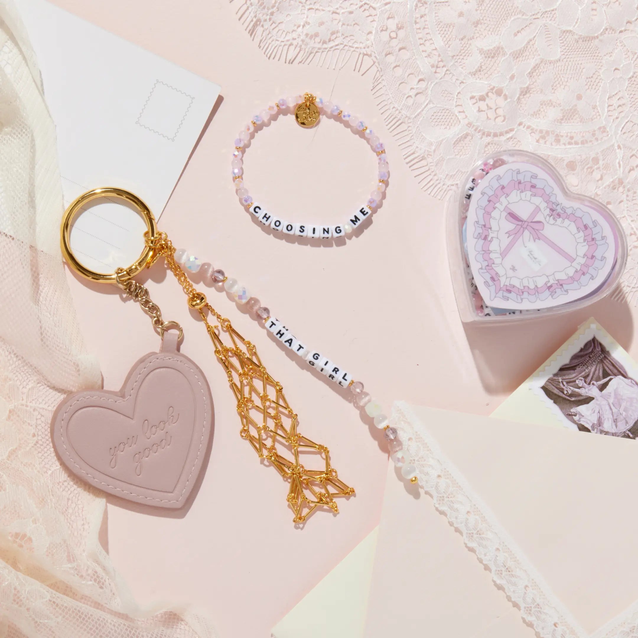 Heart-shaped keychain with gold tassel, beaded bracelet, and heart-shaped box on a textured pink surface.