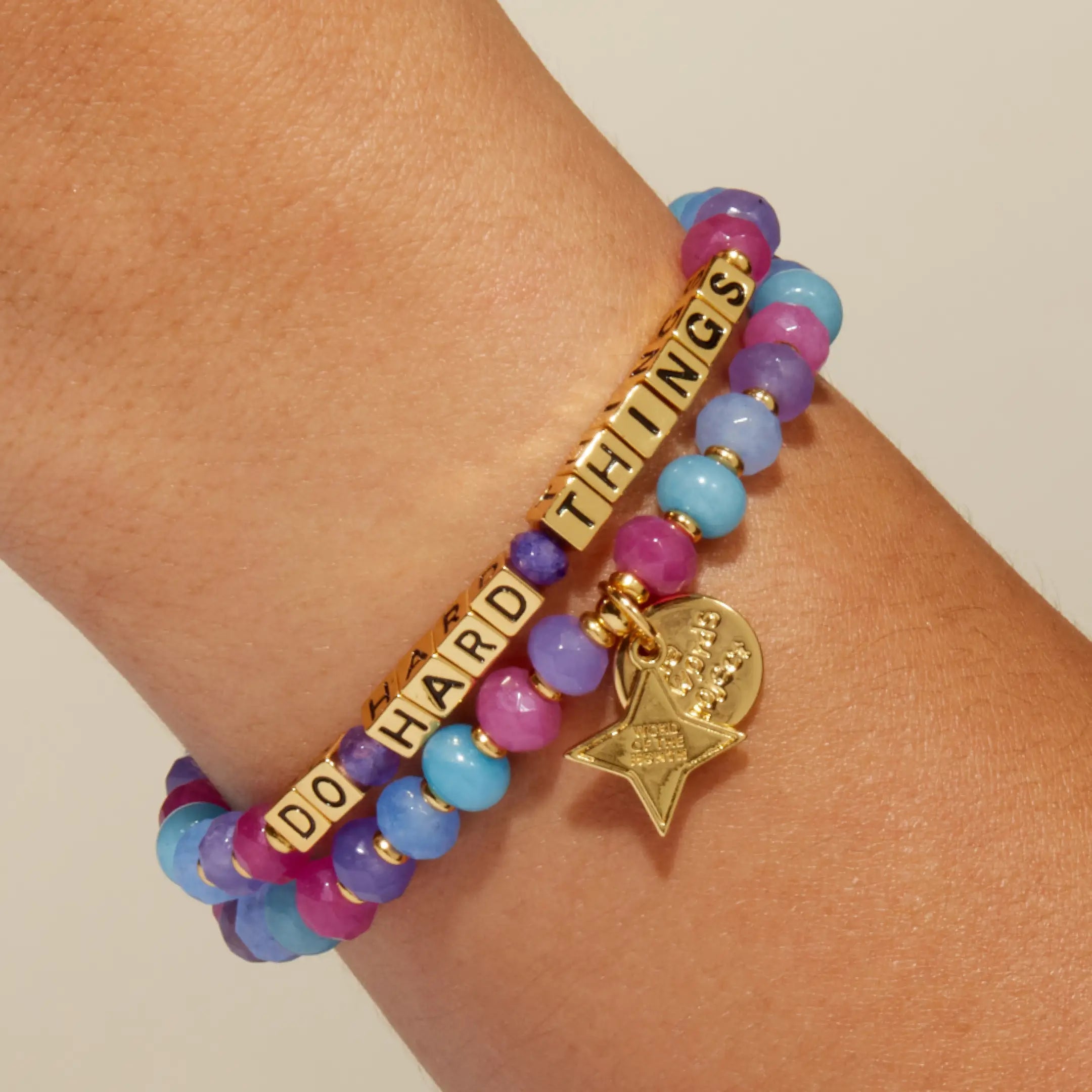 Do Hard Things September Word of the Month bracelet with gold letter beads worn on a wrist and stacked with a matching pink, purple and blue beaded bracelet