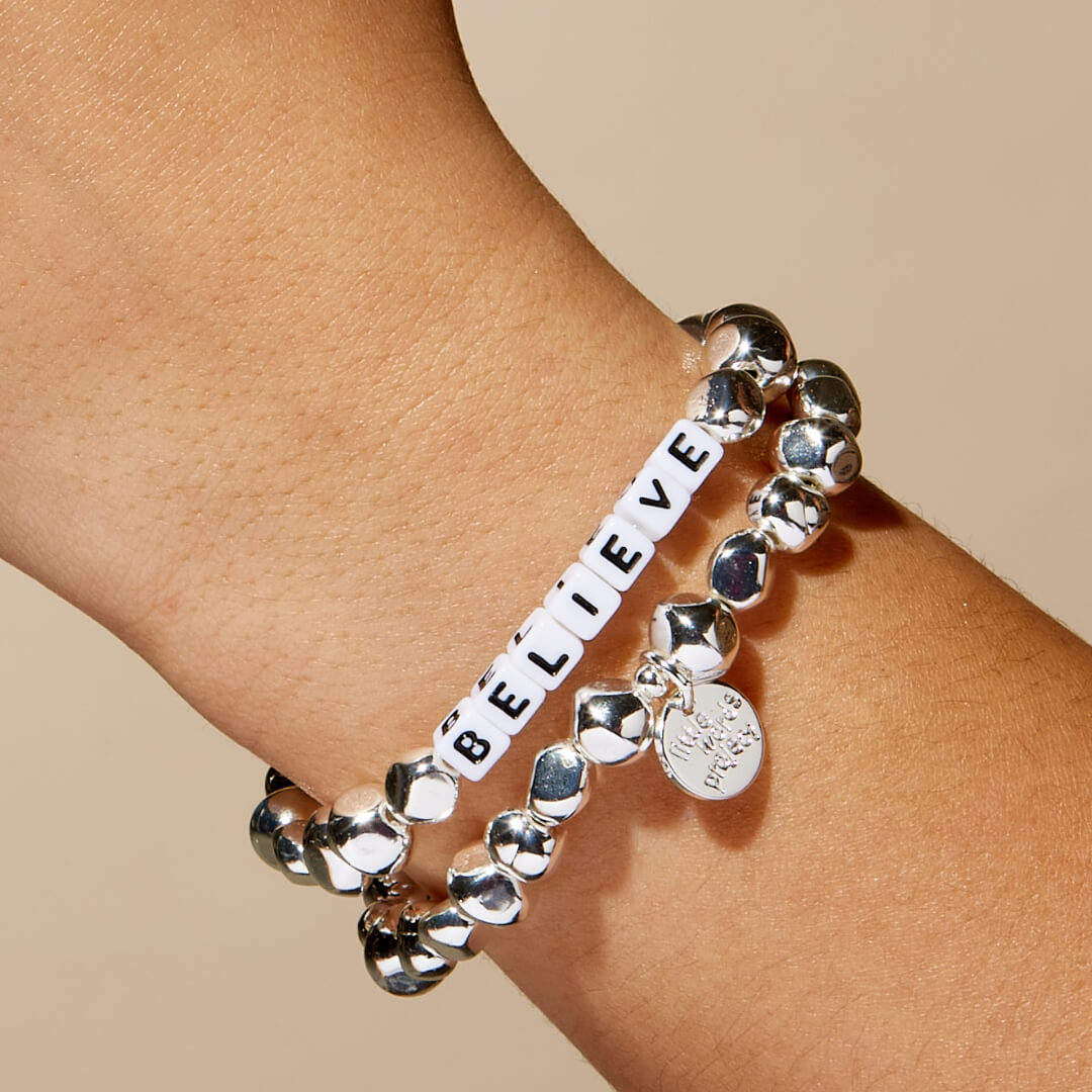 Silver bracelet with 'BELIEVE' charm on a wrist against a beige background