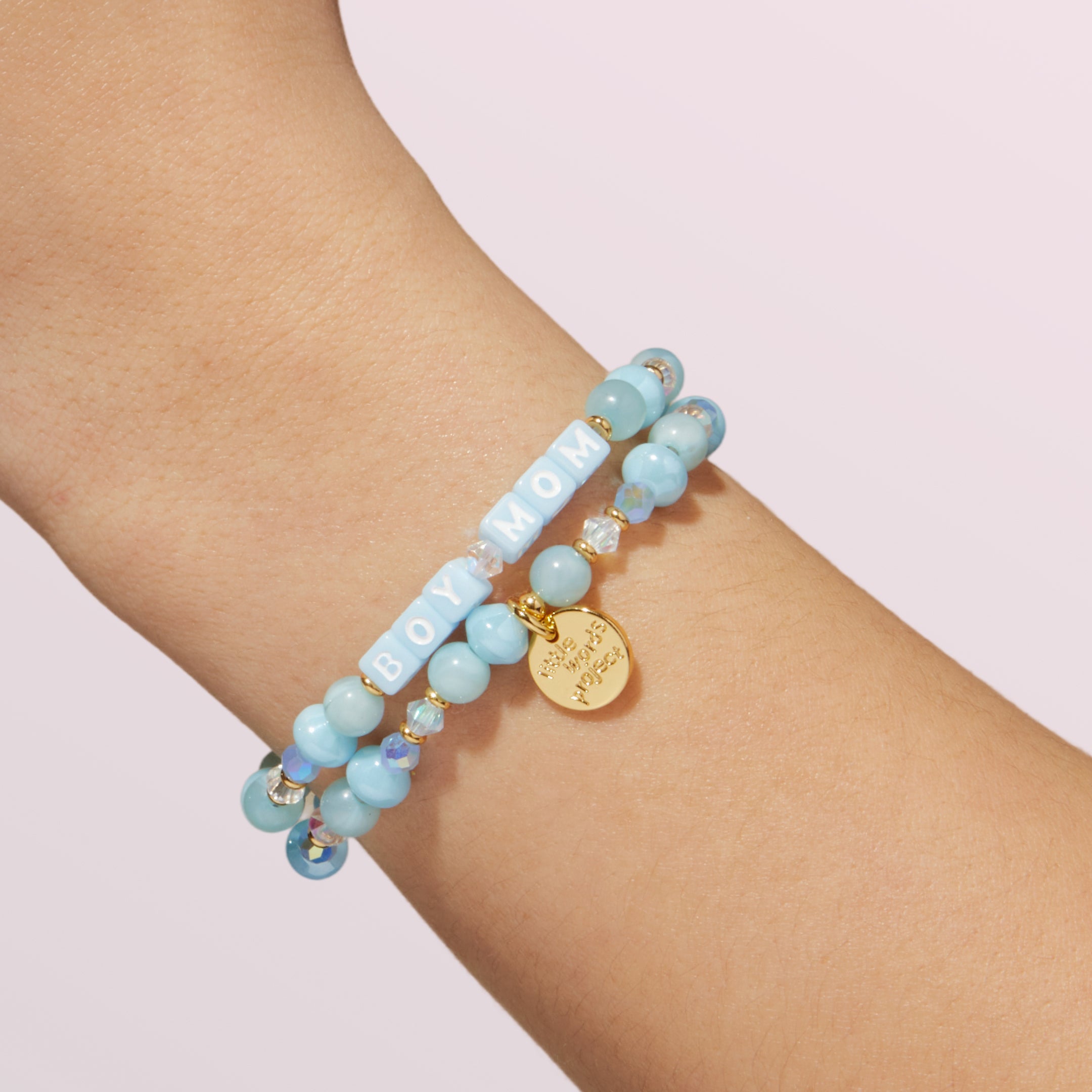 Light blue beaded bracelet with gold tag on a wrist against a light pink background