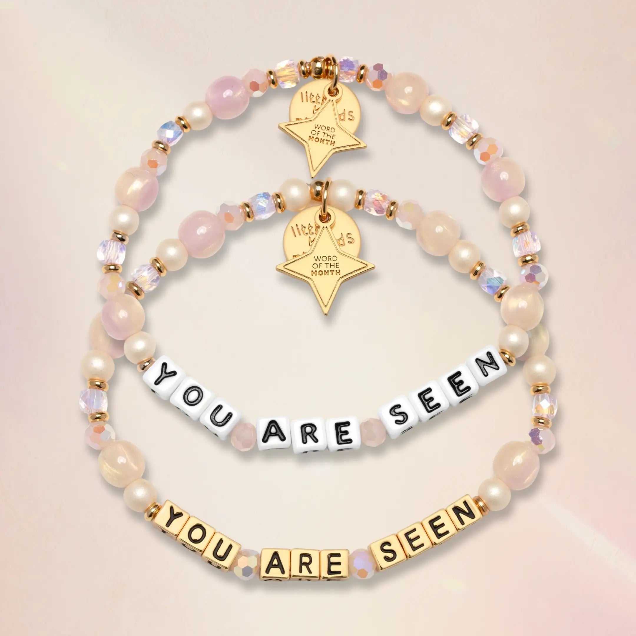 Two You Are Seen Word of the Month bracelets shown together, one with white letter beads and one with gold letter beads, on a soft pink background