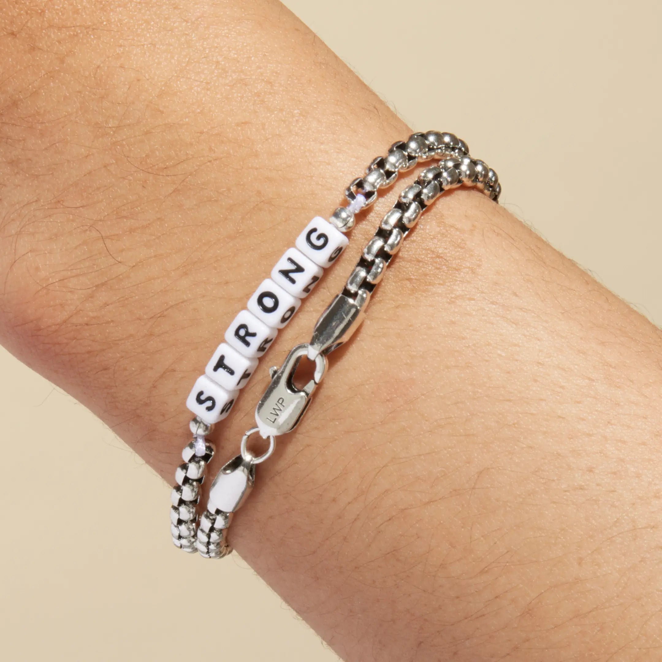 Silver box chain bracelet with white square letter beads spelling 'STRONG' worn on a wrist, stacked with a plain silver box chain bracelet. LWP lobster clasp visible in the foreground.