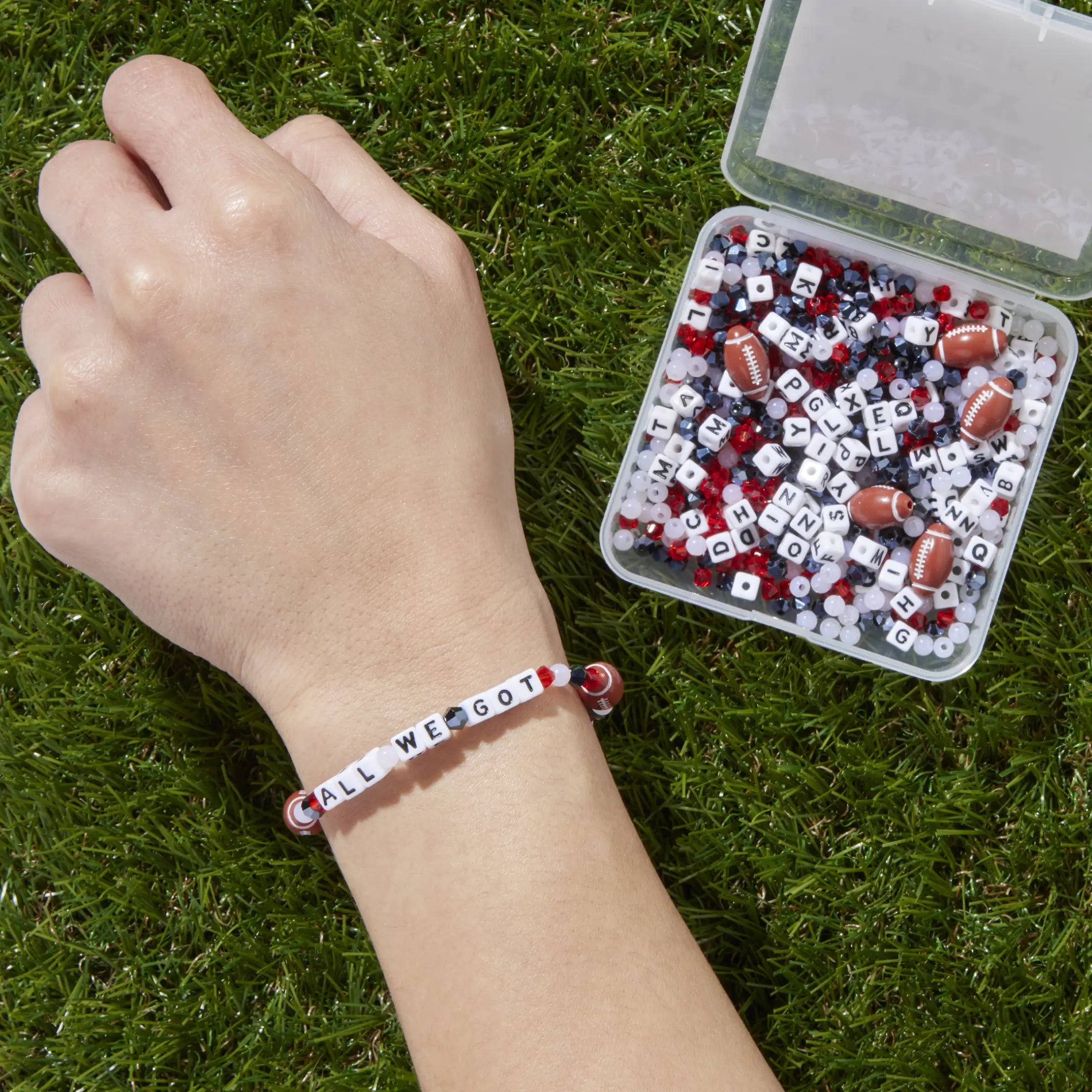 Hand wearing a bracelet with letter beads on grass next to a container of letter beads