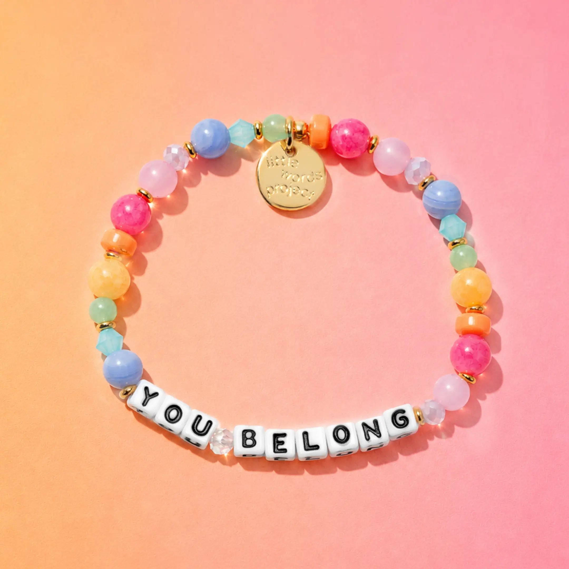 Colorful beaded bracelet with a gold tag and letter beads spelling 'YOU BELONG' on a gradient background.