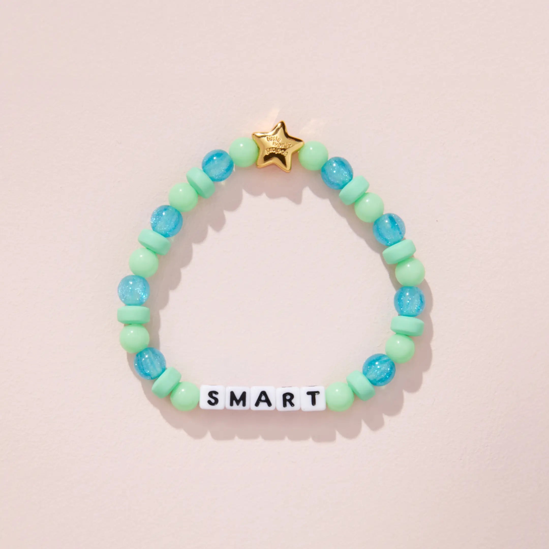 Bracelet with green and blue beads and a gold tag on a light pink background