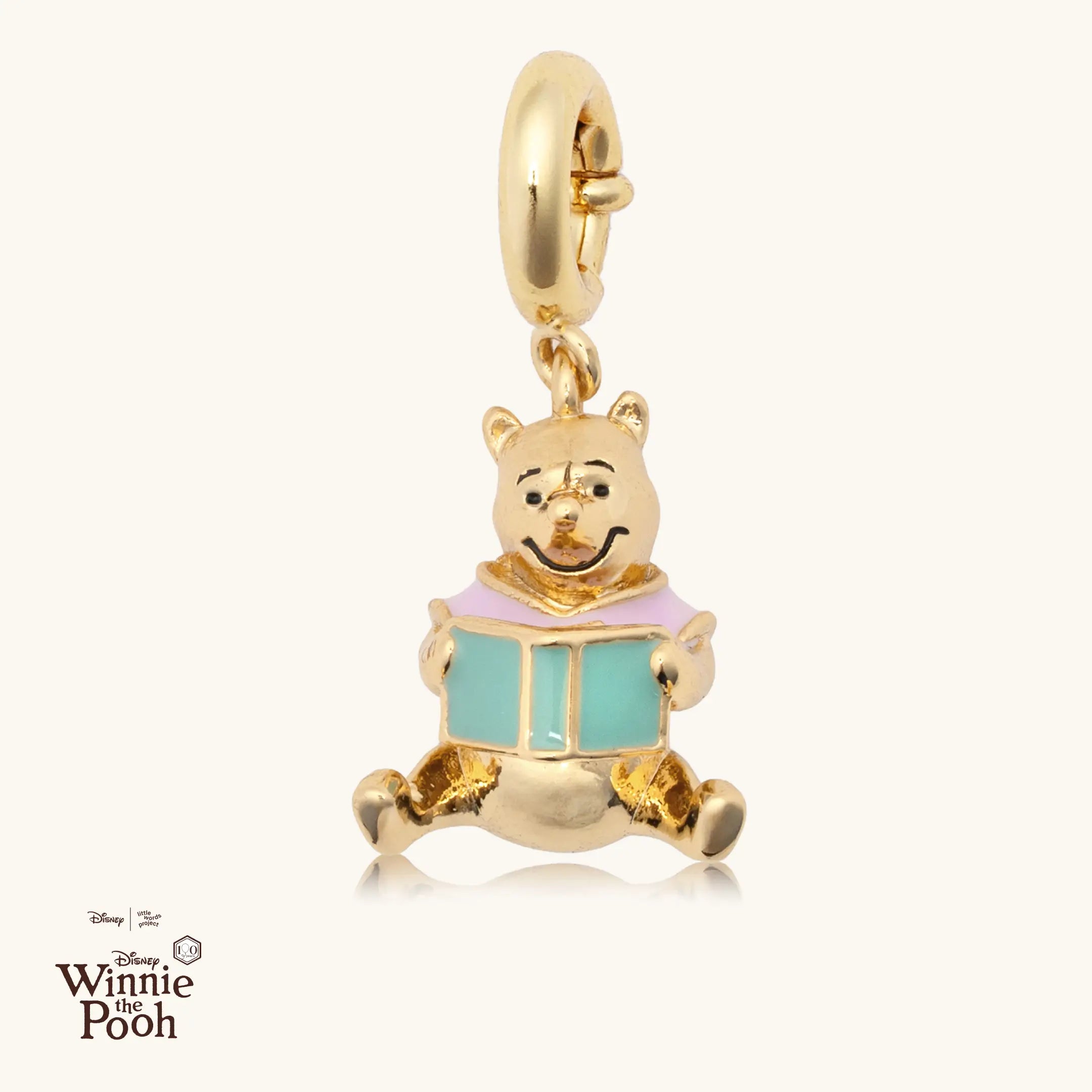 Gold charm of a cartoon bear holding a blue book on a white background, with Disney Winnie the Pooh logo.