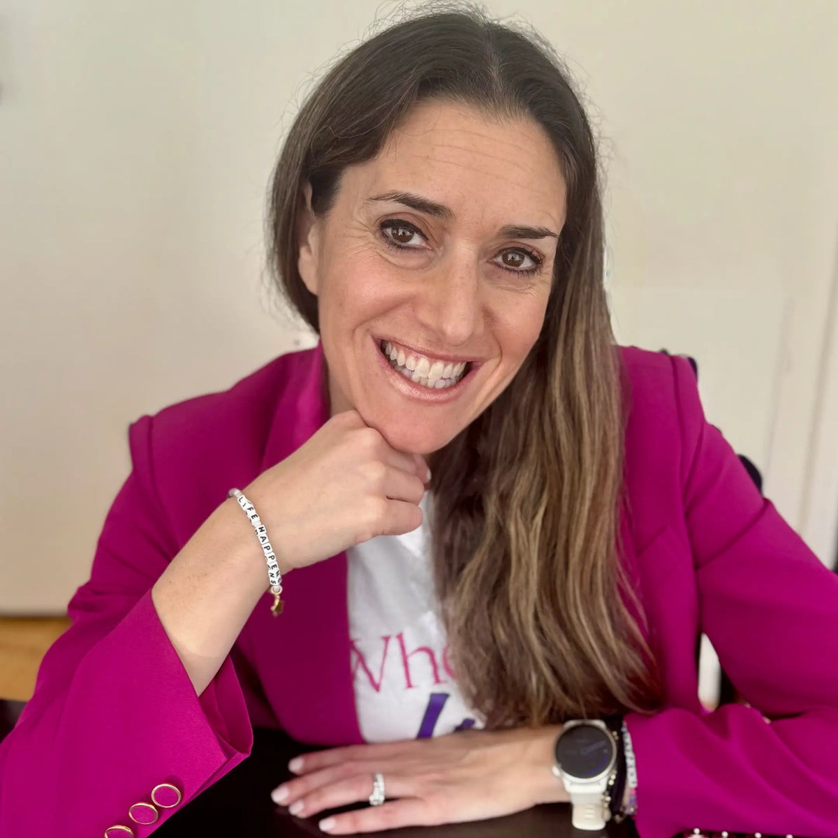 A woman with long brown hair smiles broadly, resting her chin on her fist. She is wearing a bright magenta blazer over a white graphic t-shirt, a watch, rings, and a beaded bracelet on her wrist — likely the "LIFE HAPPENS" bracelet from the previous images. She is seated at a table in a bright, neutral indoor setting.