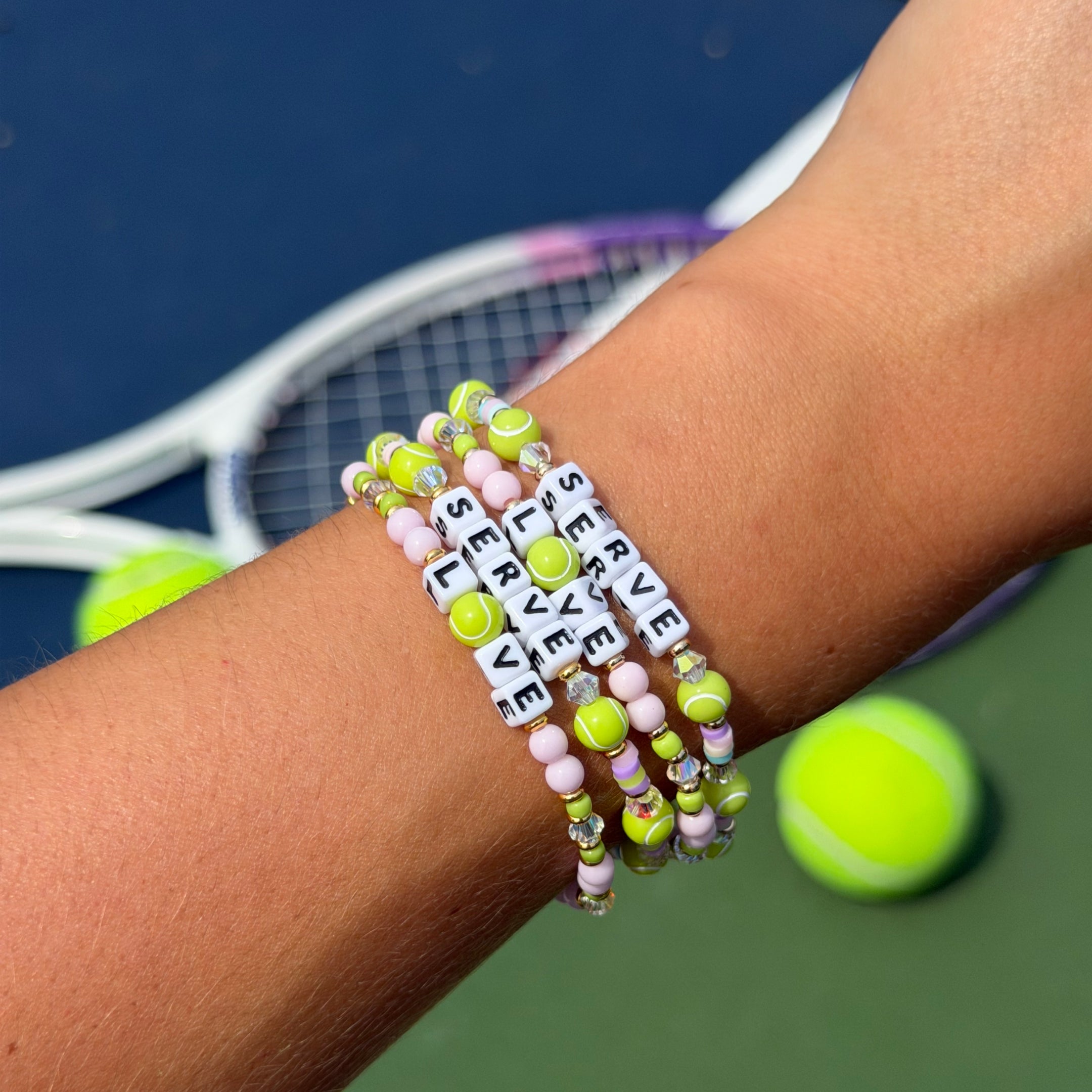 A close-up of a person's wrist wearing four stacked beaded bracelets with tennis-themed words, including neon yellow-green tennis balls, and pastel pink beads. A tennis racket and tennis balls are blurred in the background on a tennis court.