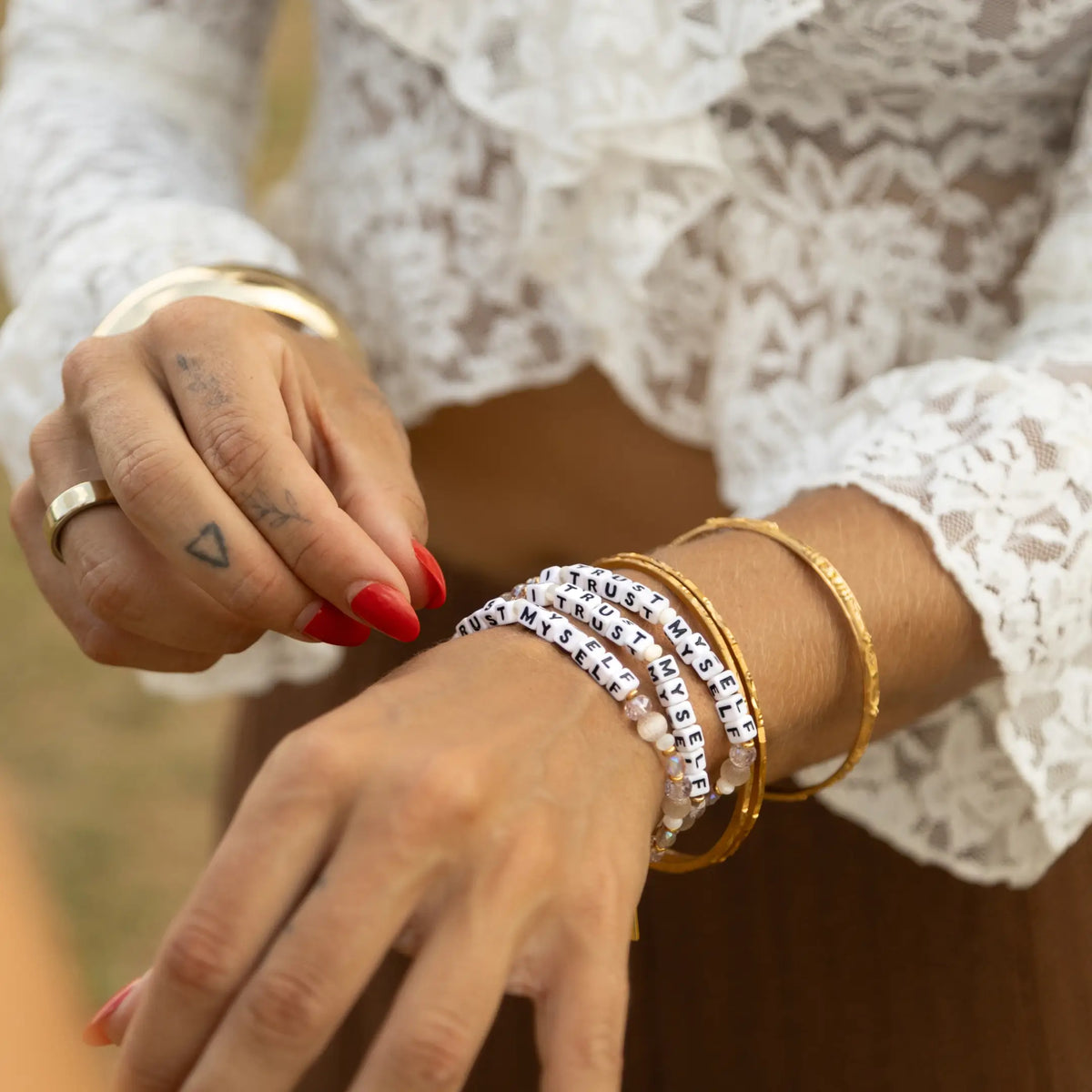 I Trust Myself Tori Deal Kindfluencer bracelets stacked with gold bangles on model's wrist, white letter beads and blush crystals, white lace sleeve