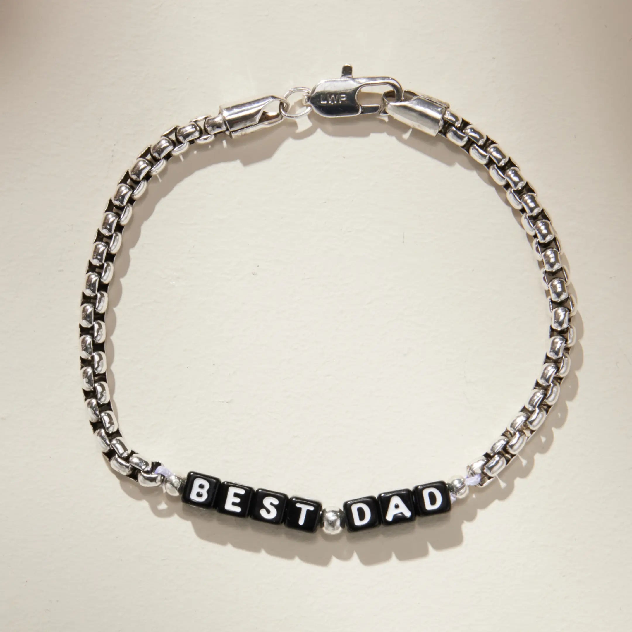 Silver box chain bracelet with black square letter beads spelling 'BEST DAD' at the center, laid flat on a cream background. Features an LWP-engraved silver lobster clasp closure.