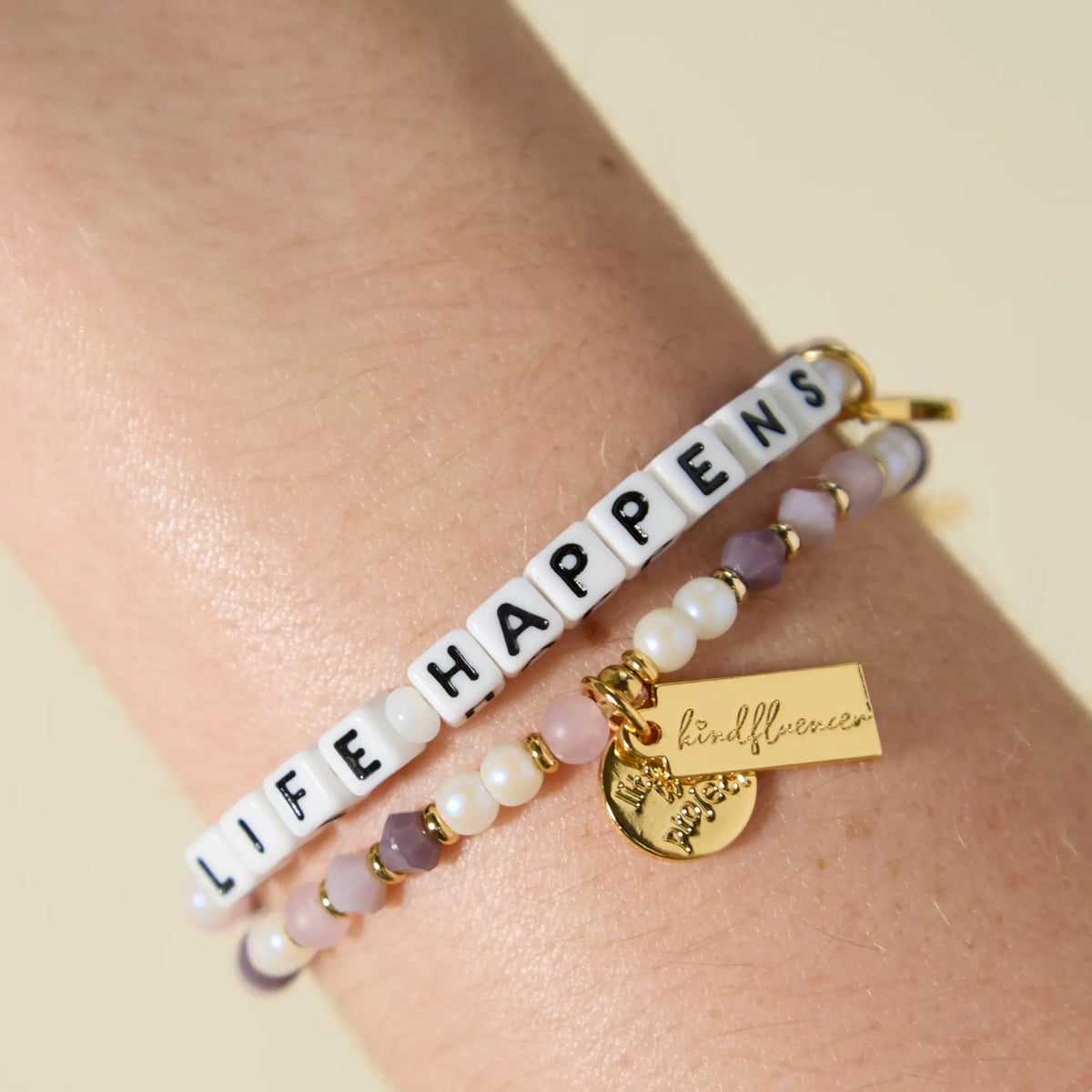 Two beaded bracelets worn on a wrist. The top bracelet spells "LIFE HAPPENS" in white square letter beads with black text. Both bracelets feature a mix of soft pink, mauve, purple, and white pearl beads with small gold spacers. A gold rectangular charm with cursive text and a small round coin charm hang from the lower bracelet, photographed against a warm, neutral background.