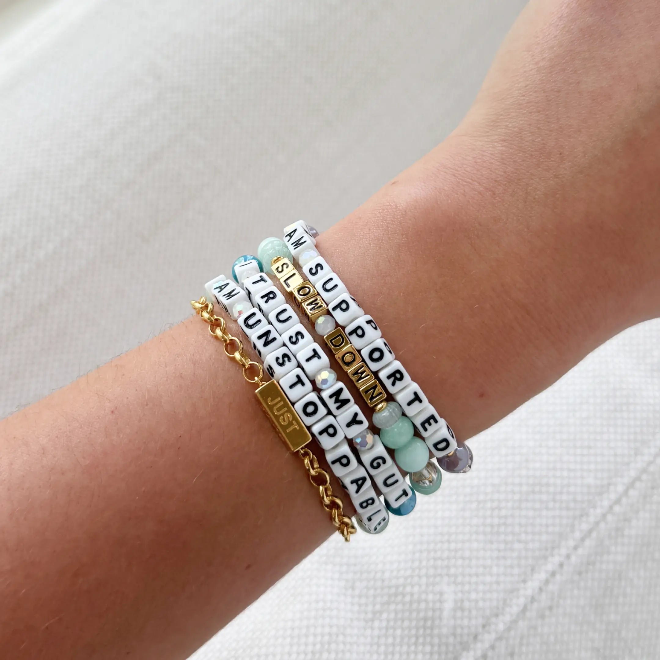 A wrist stacked with multiple beaded bracelets reading "Supported," "Slow Down," "Trust My Gut," "Unstoppable," and a gold chain bracelet with a "Just" bar charm.