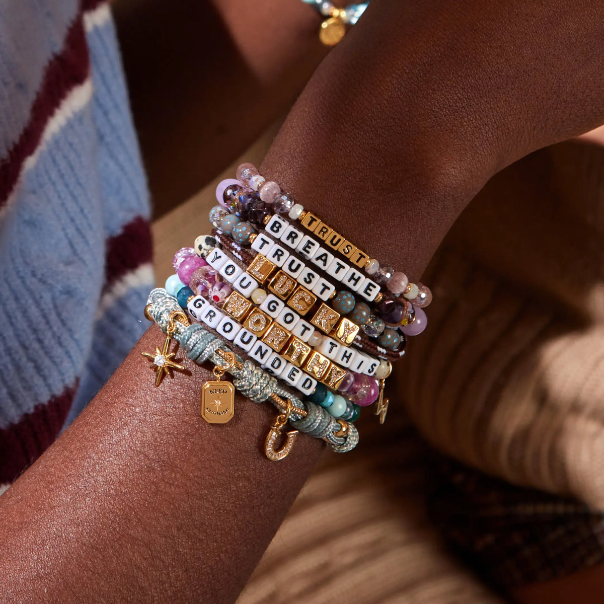 Stack of seven fall bracelets worn on a wrist spelling Breathe, Trust, Trust, Lucky, You Got This, Worthy, and Grounded, in beaded, CZ letter block, and paracord styles