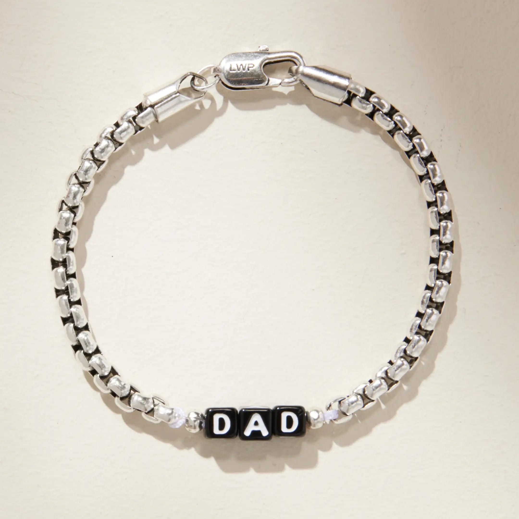 Silver box chain bracelet with black square letter beads spelling 'DAD' at the center, laid flat on a cream background. Features an LWP-engraved lobster clasp closure