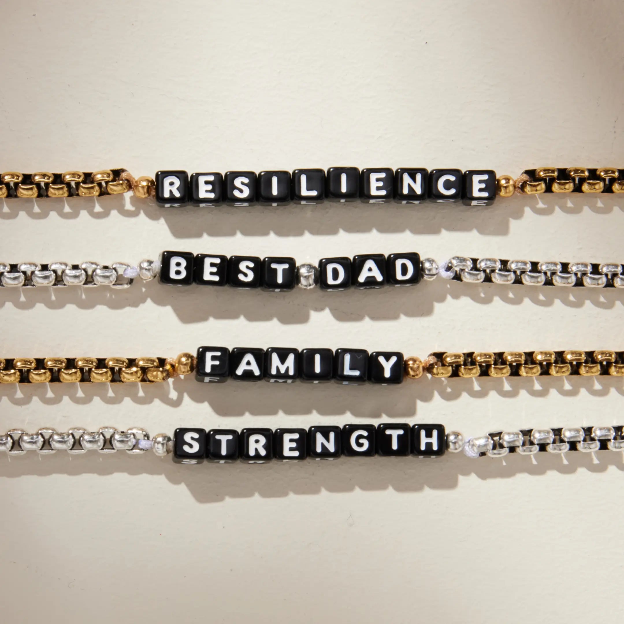 Four box chain bracelets laid horizontally on a cream background, each featuring black square letter beads spelling different words. From top to bottom: 'RESILIENCE' on a gold chain, 'BEST DAD' on a silver chain, 'FAMILY' on a gold chain, and 'STRENGTH' on a silver chain.