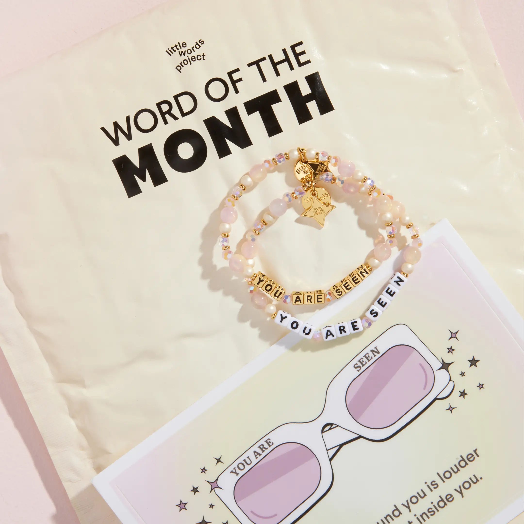 You Are Seen Word of the Month bracelets in white and gold letter beads resting on a Little Words Project Word of the Month mailer with a sunglasses insert card