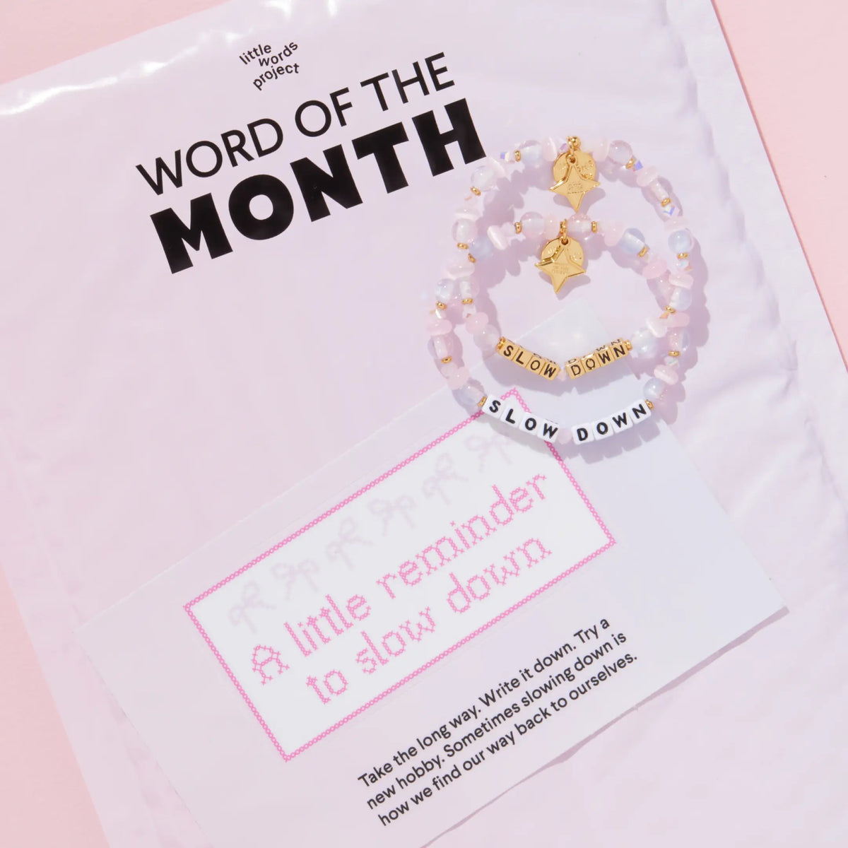 Slow Down October Word of the Month bracelets in white and gold letter beads on a pink Word of the Month mailer with A little reminder to slow down sticker, packaging on pink background