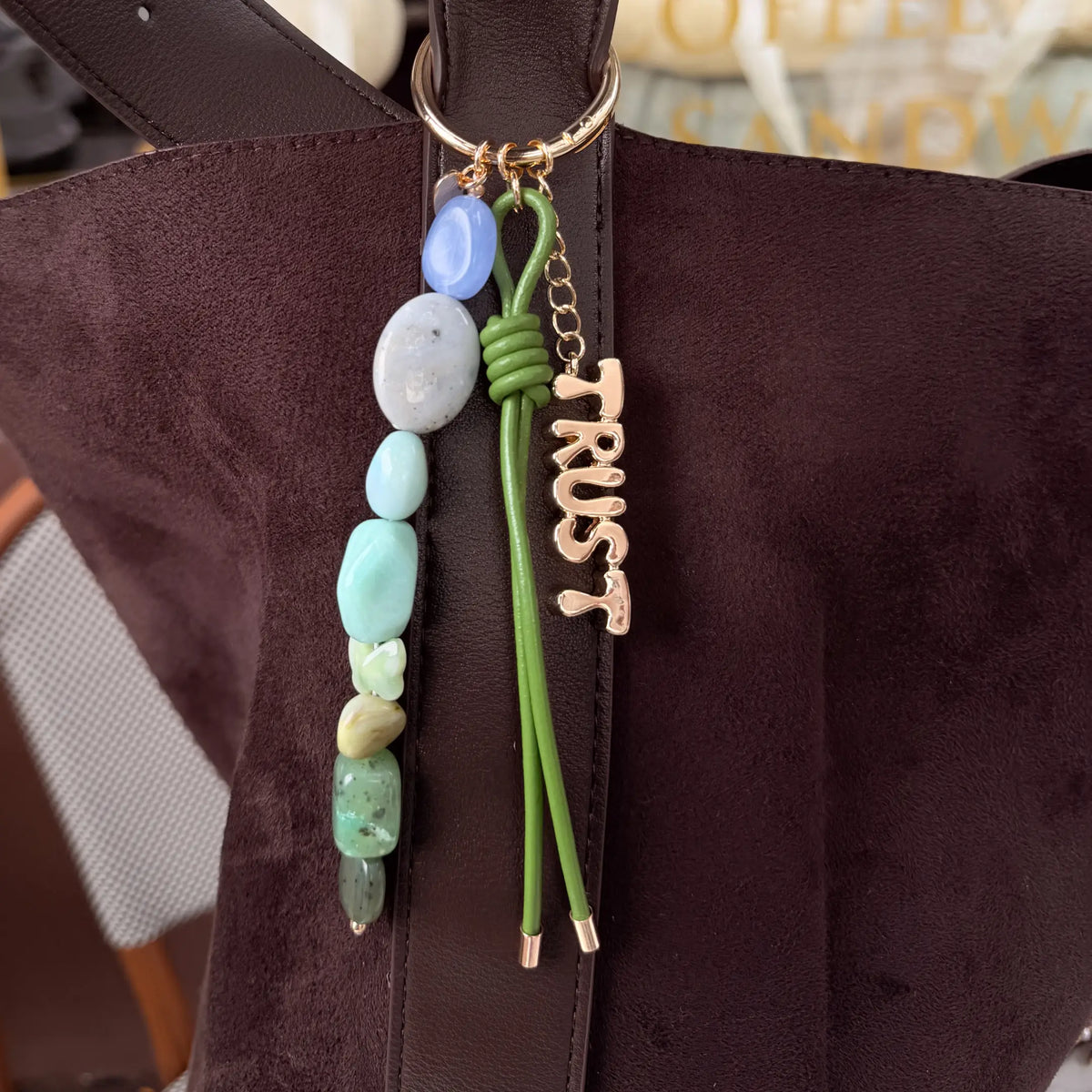 Trust Bag Charm clipped to a brown suede bag, blue and green stone beaded strand, green knotted cord, and gold Trust charm, styled lifestyle shot