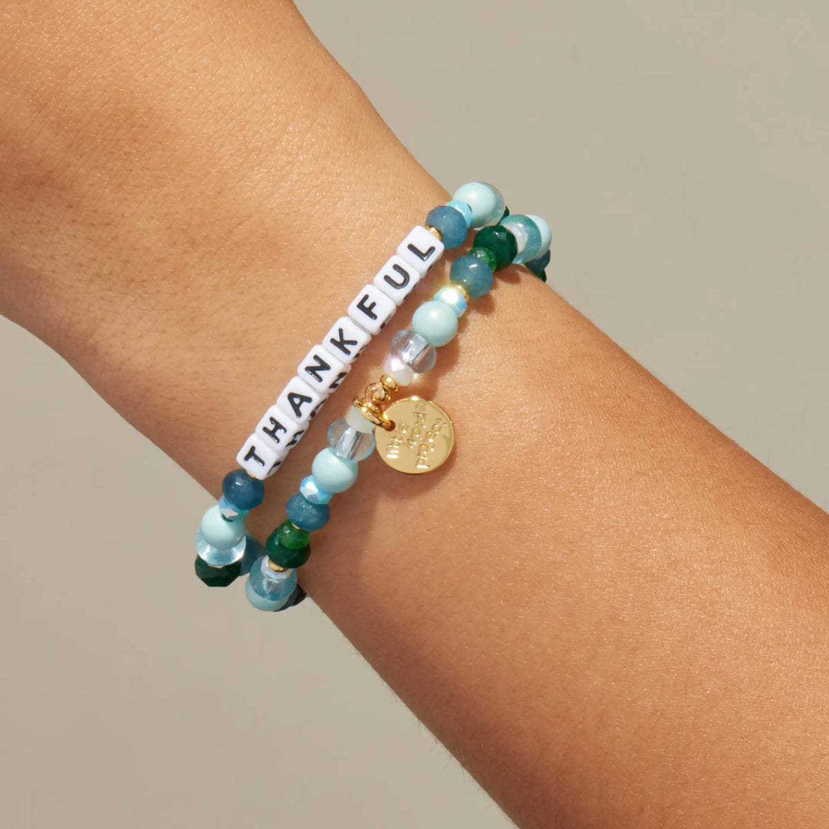 Custom Rebel Cozy bracelet worn on a wrist, white letter beads spelling Thankful with light blue pearls and emerald green crystals