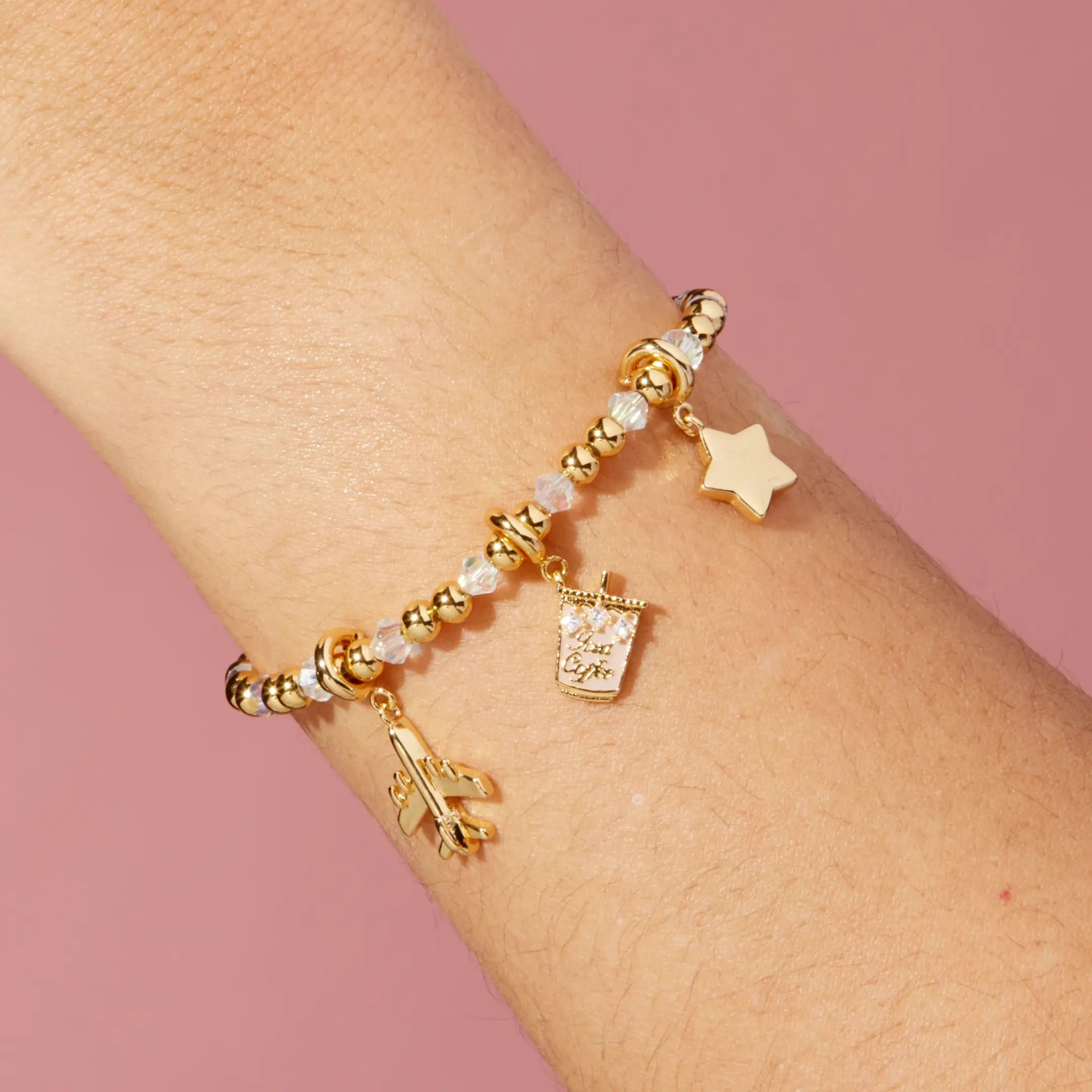 Gold bracelet with star, iced coffee, and airplane charms on a wrist against a pink background