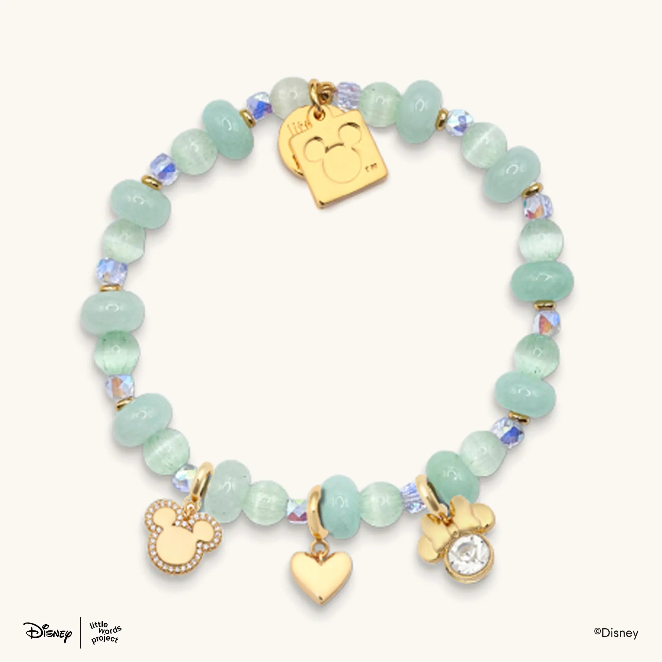 Mickey Mouse and Minnie Mouse Charm Bracelet from Disney Mickey and Friends, flat lay on cream with mint green beads, a gold plated casted Mickey Mouse tag charm, a Minnie Mouse bow charm, a heart charm and a crystal charm