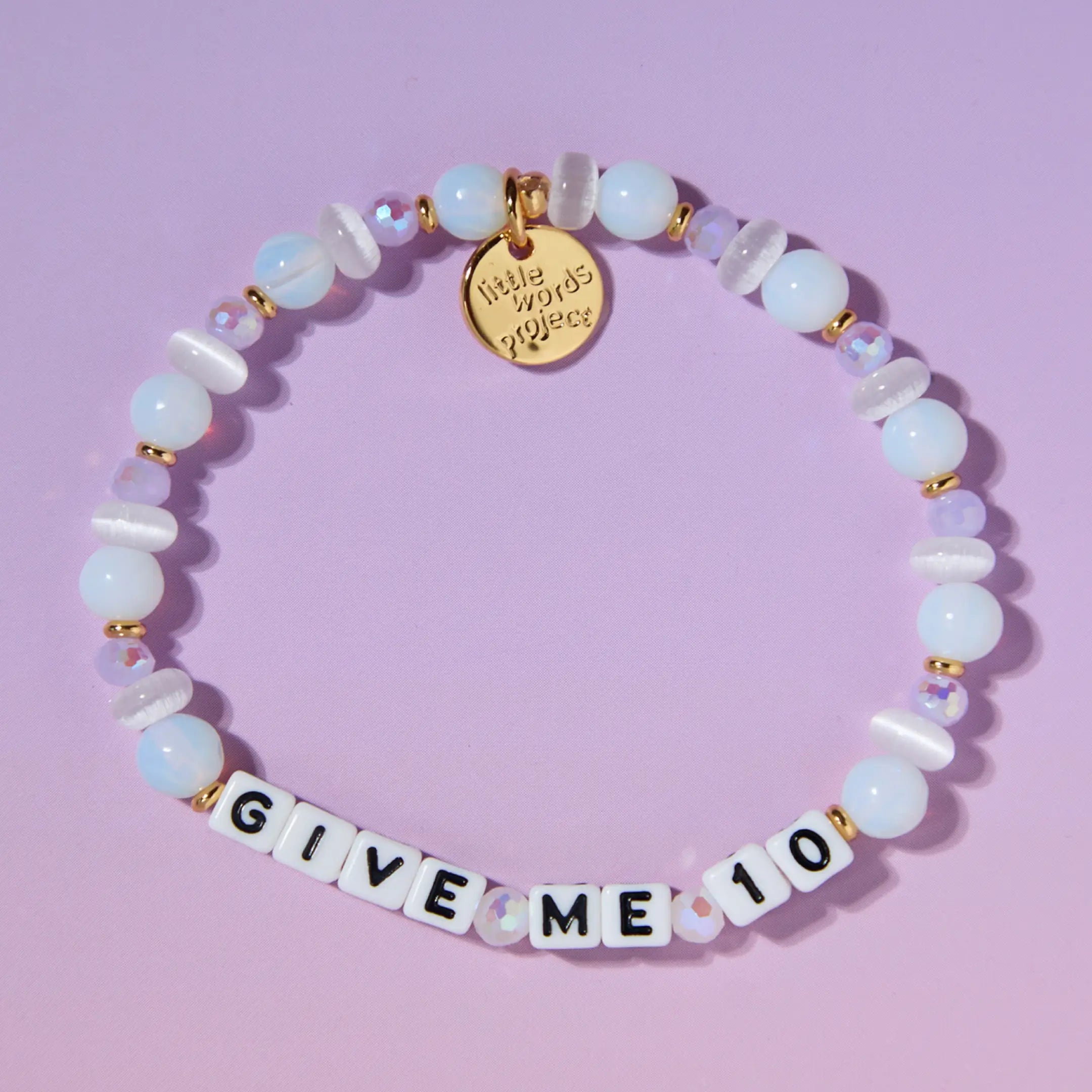 A beaded stretch bracelet arranged in a circle on a light purple background. The bracelet features white and pale blue beads, small gold spacer beads and white letter tiles spelling 'GIVE ME 10'.