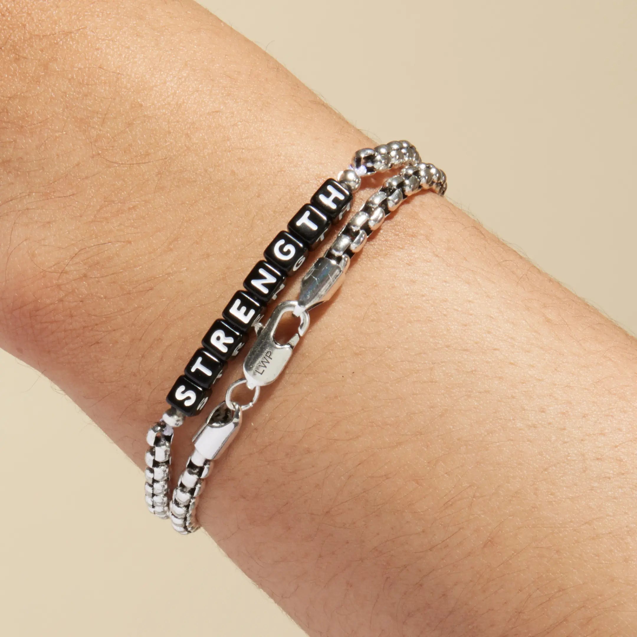 Silver box chain bracelet with black square letter beads spelling 'STRENGTH' worn on a wrist, stacked with a plain silver box chain bracelet. LWP-engraved silver lobster clasp visible in the foreground.