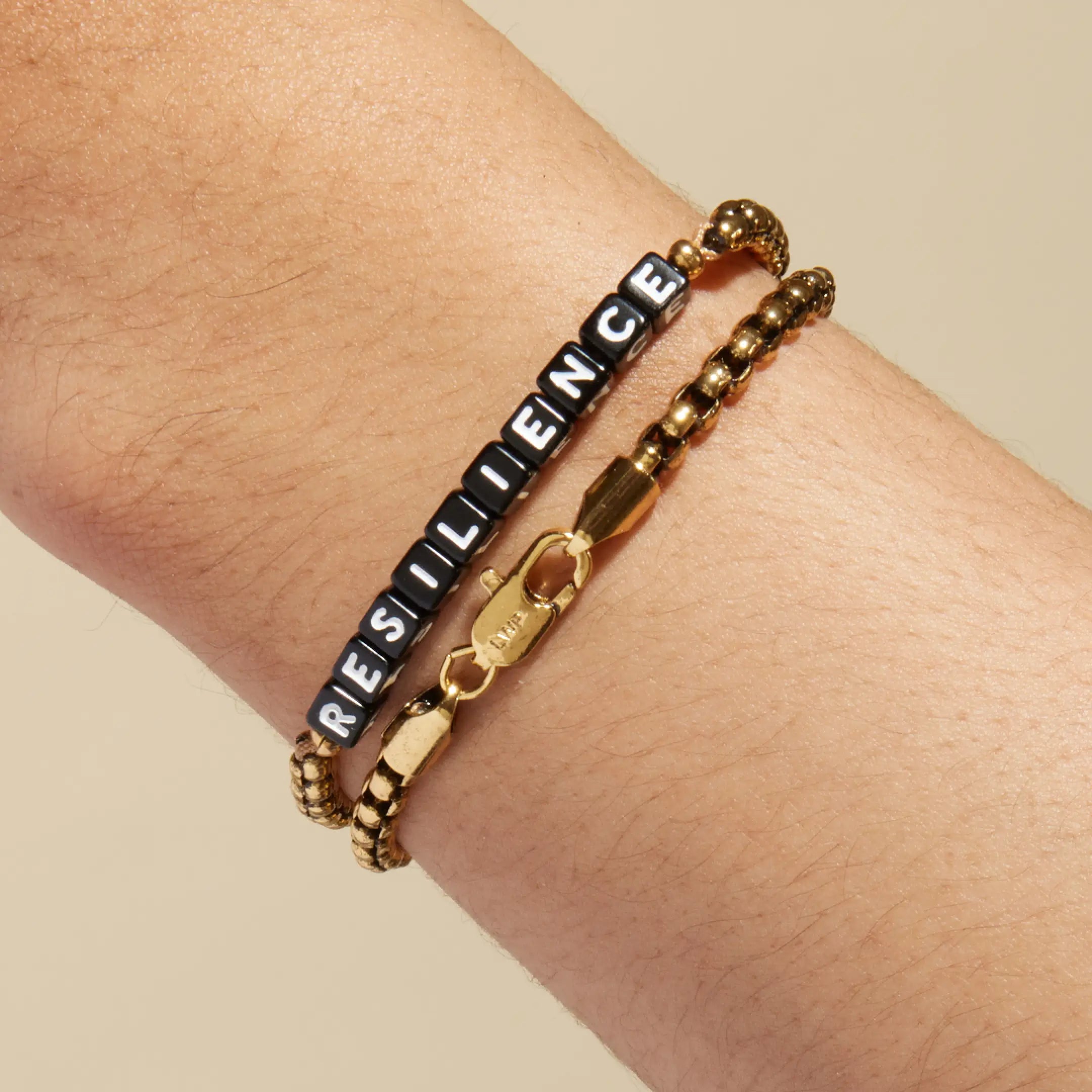 Gold chain bracelet with black square letter beads spelling 'RESILIENCE' worn on a wrist, stacked with a plain silver box chain bracelet. LWP-engraved gold lobster clasp visible in the foreground.
