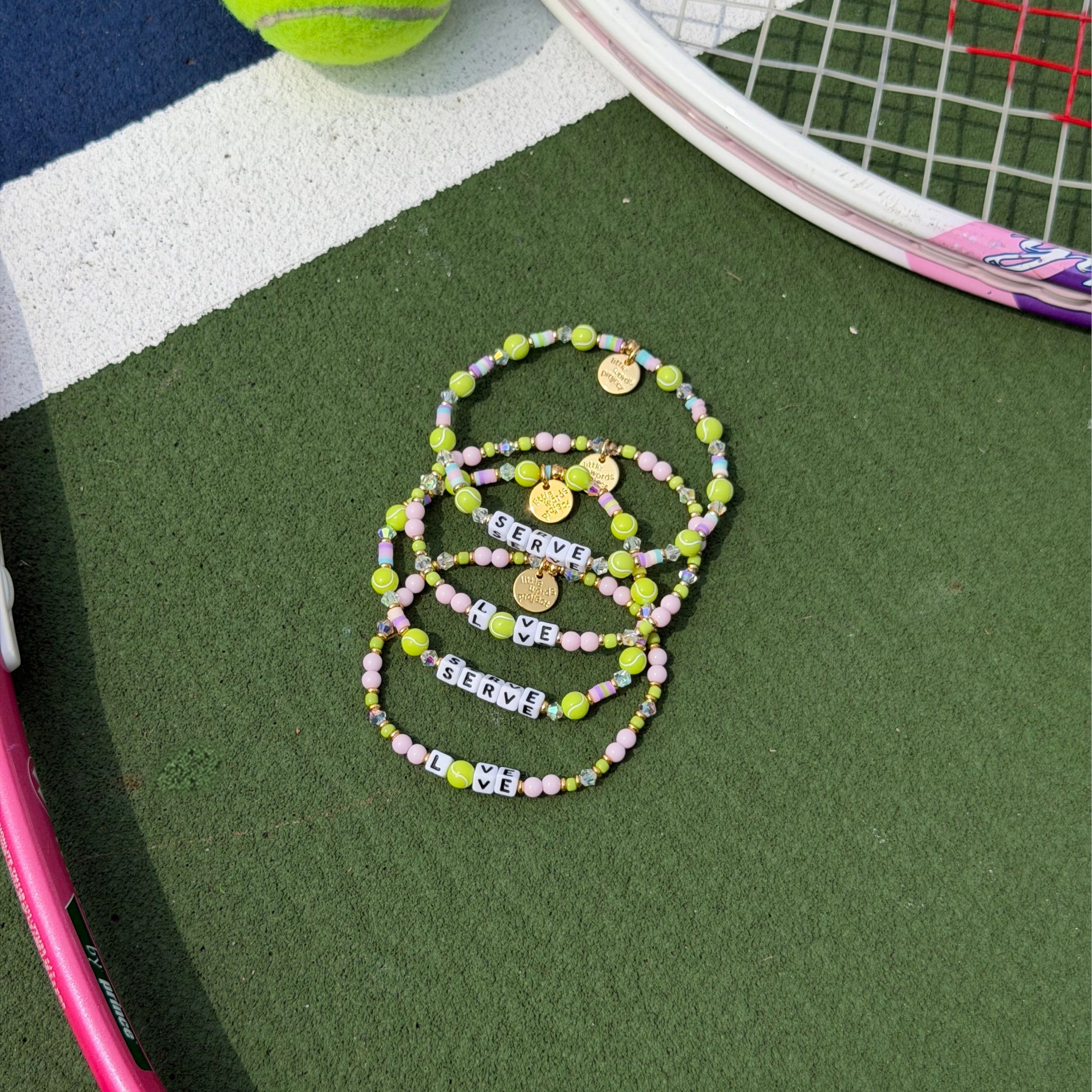 Four beaded bracelets arranged on a green tennis court with a tennis racket and ball partially visible.