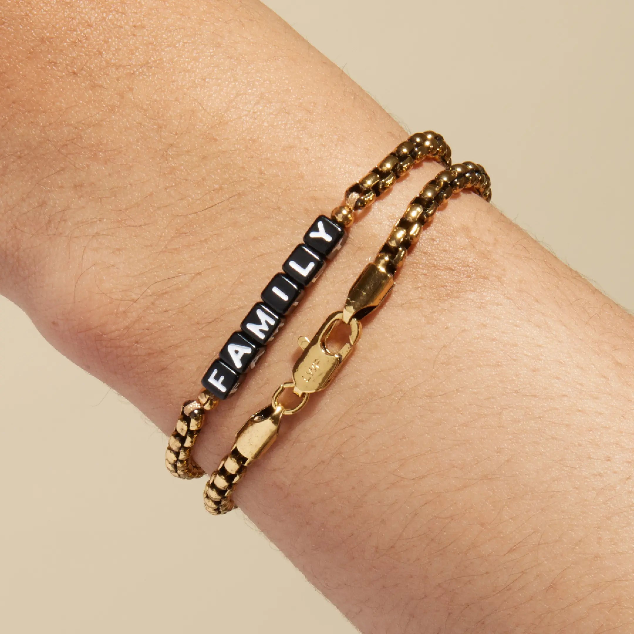 Gold box chain bracelet with black square letter beads spelling 'FAMILY' worn on a wrist, stacked with a plain gold box chain bracelet. LWP-engraved gold lobster clasp visible in the foreground.