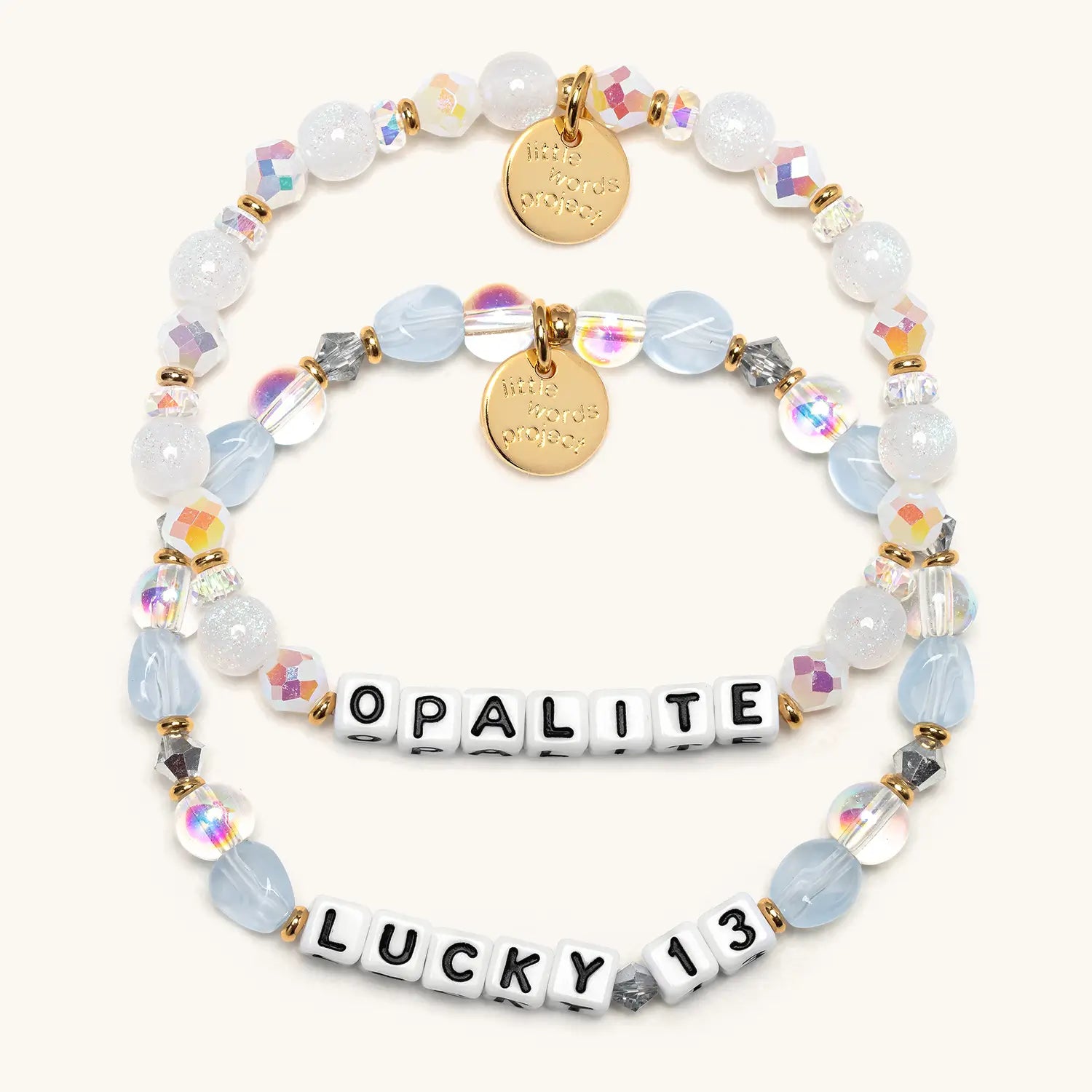 Flat-lay of two stacked bracelets: a white iridescent Opalite bracelet and a frosted-blue Lucky 13 bracelet, both with gold charms and white OPALITE and LUCKY 13 letter beads