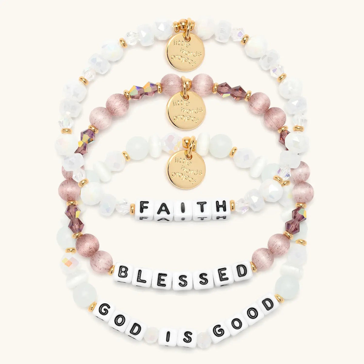 Faith Bracelet Set stacked flat-lay showing the Faith bracelet in white and clear crystal beads, the Blessed bracelet in mauve cat eye and purple crystals, and the God is Good bracelet in white cat eye and frosted beads, all with white letter beads, on cream background