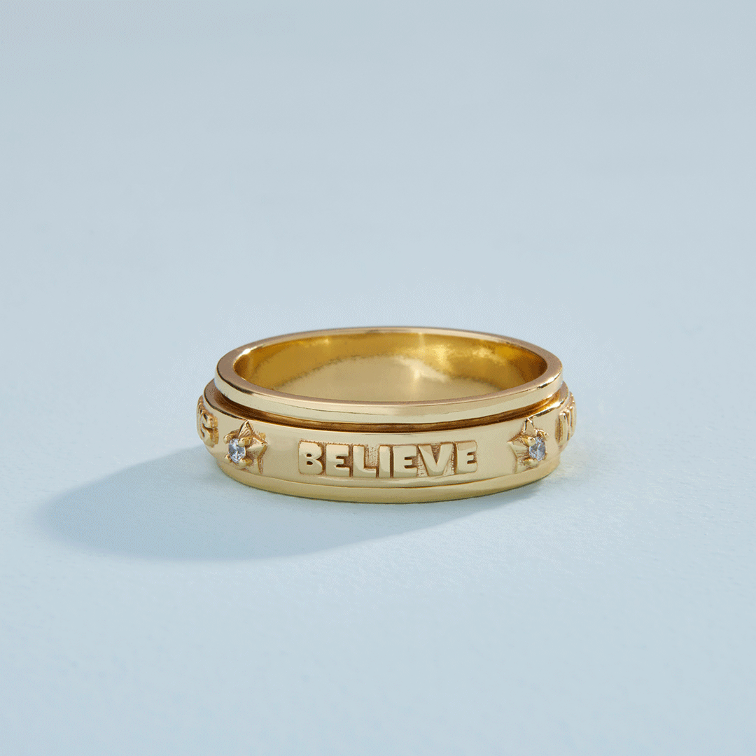 Gold "BELIEVE" spinner ring by Little Words Project, designed to spin, on a light blue background.
