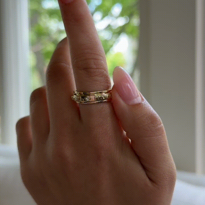 Hand wearing a gold spinner ring with engraved text, held up near a window.