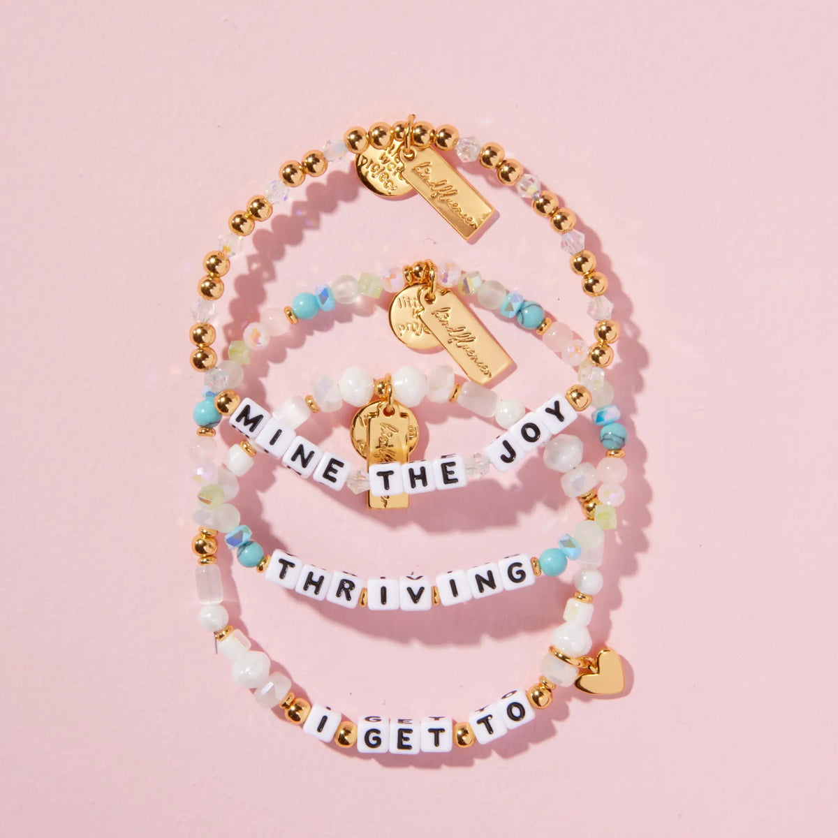 Breast Cancer Awareness Collective stack flat-lay on pink background, the Mine the Joy bracelet with gold plated beads and clear crystals, the Thriving bracelet with turquoise, pink, and sea green beads, and the I Get To bracelet with white faux pearls and a gold heart charm