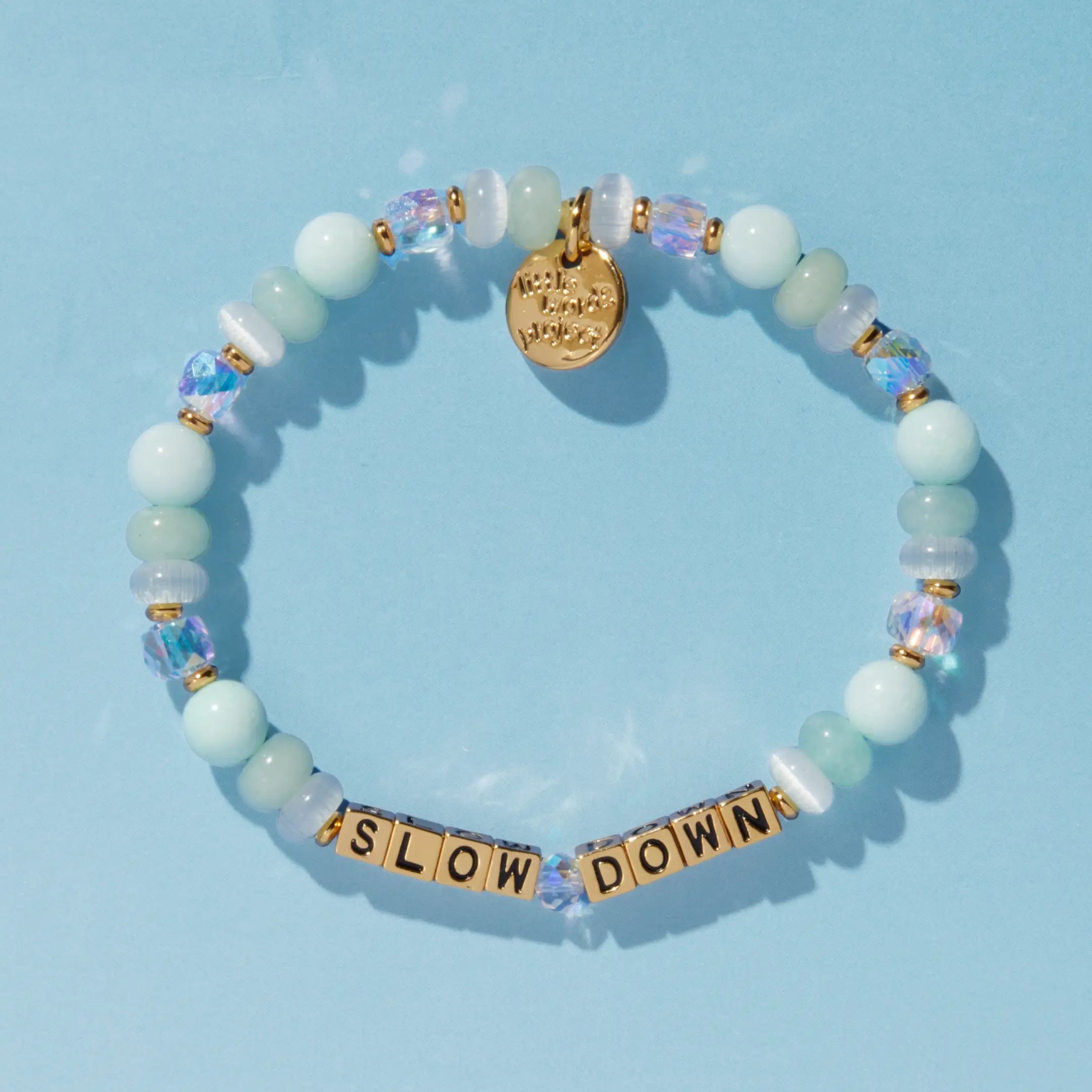 A beaded bracelet spelling "Slow Down" in gold letter cubes, with mint green, opalescent, and iridescent crystal beads and a gold Little Words Project charm, on a blue background.