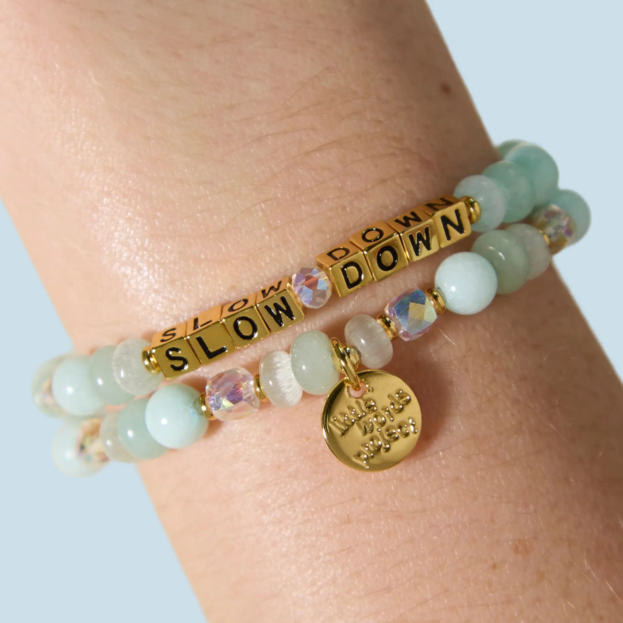 Two bracelets stacked on a wrist — one with gold letter beads reading "Slow Down" and one with mint green, iridescent crystal, and white beads featuring a gold Little Words Project charm pendant.