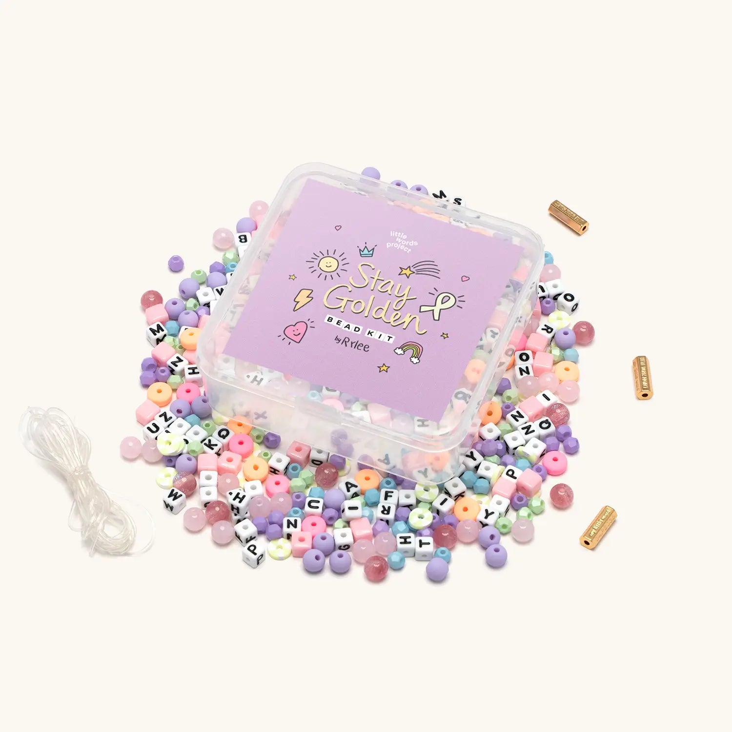 Stay Golden Bead Kit Rylee Ta with purple box lid and loose letter beads, pastel star and heart beads and elastic cord spilling out, flat lay on cream background
