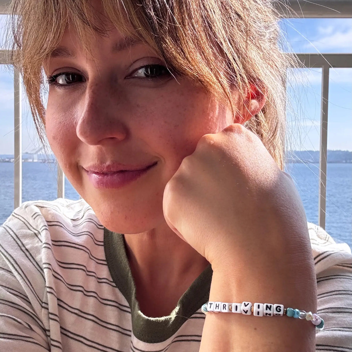 Katie Thurston wearing her Thriving bracelet, on-model selfie with her chin resting on her hand by the water