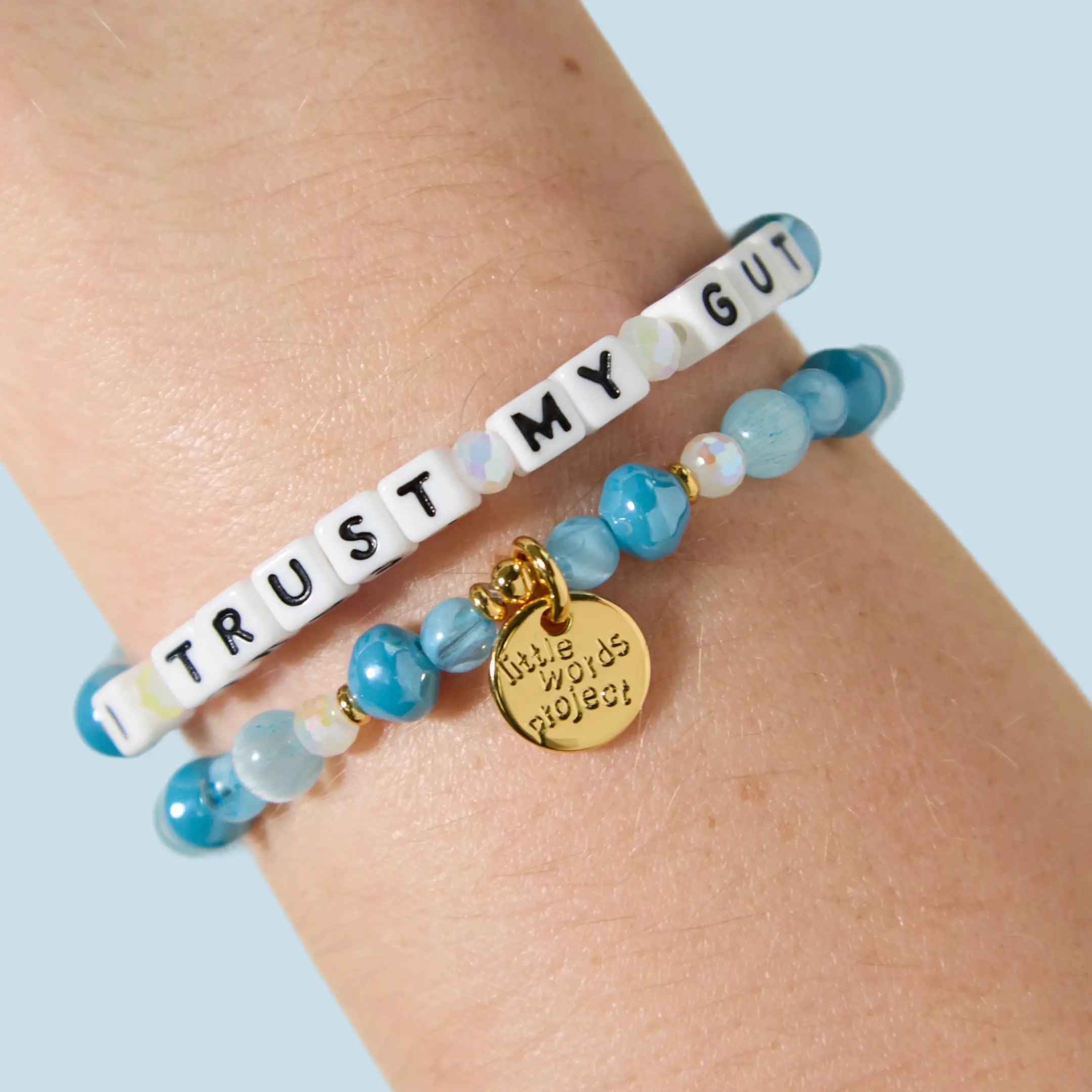 Two bracelets stacked on a wrist — one with white letter beads reading "I Trust My Gut" and one with blue agate, aqua, and opalescent beads featuring a gold Little Words Project charm pendant.