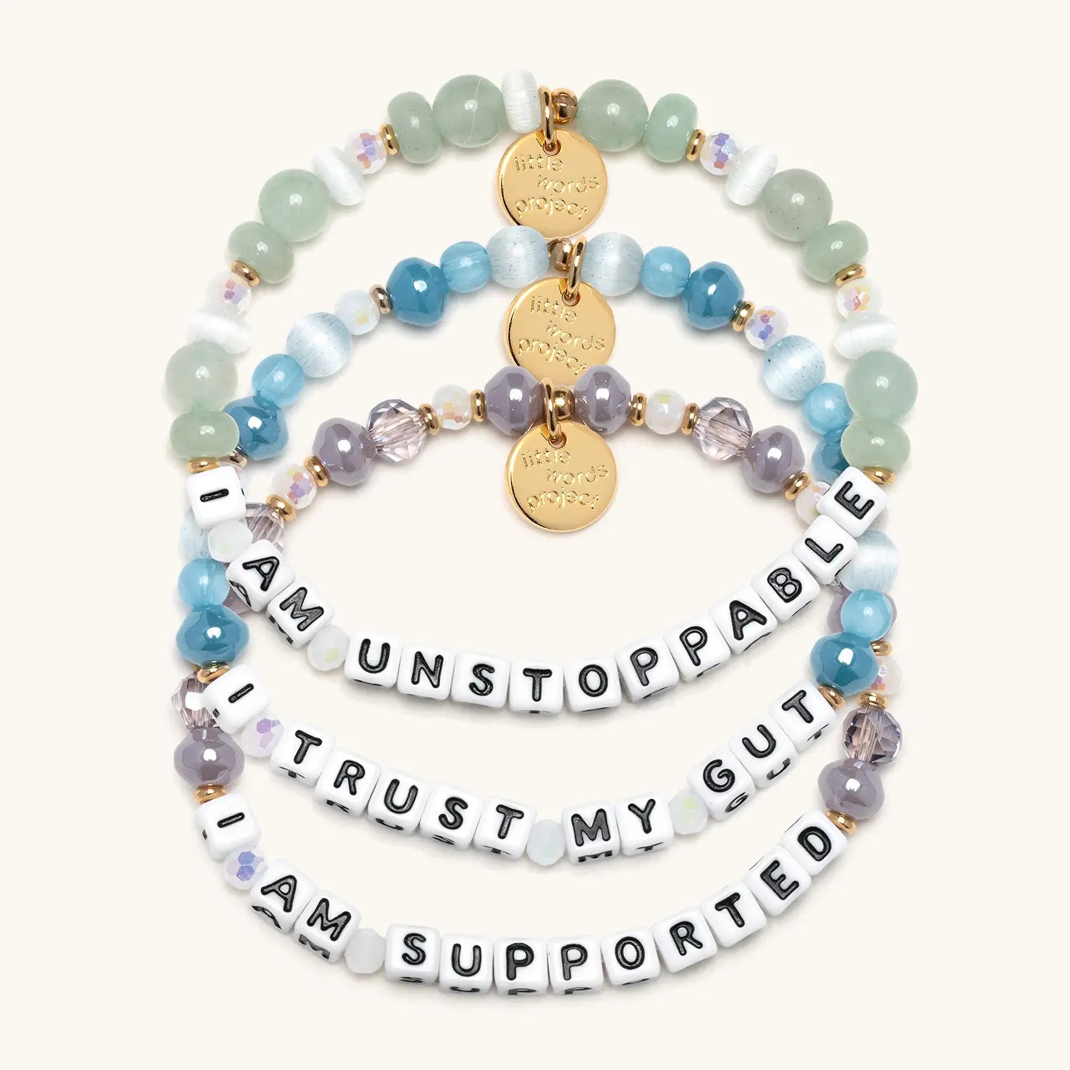 Three stacked beaded bracelets reading "I Am Unstoppable," "Trust My Gut," and "I Am Supported" in blue, green, and purple tones with gold coin charms.