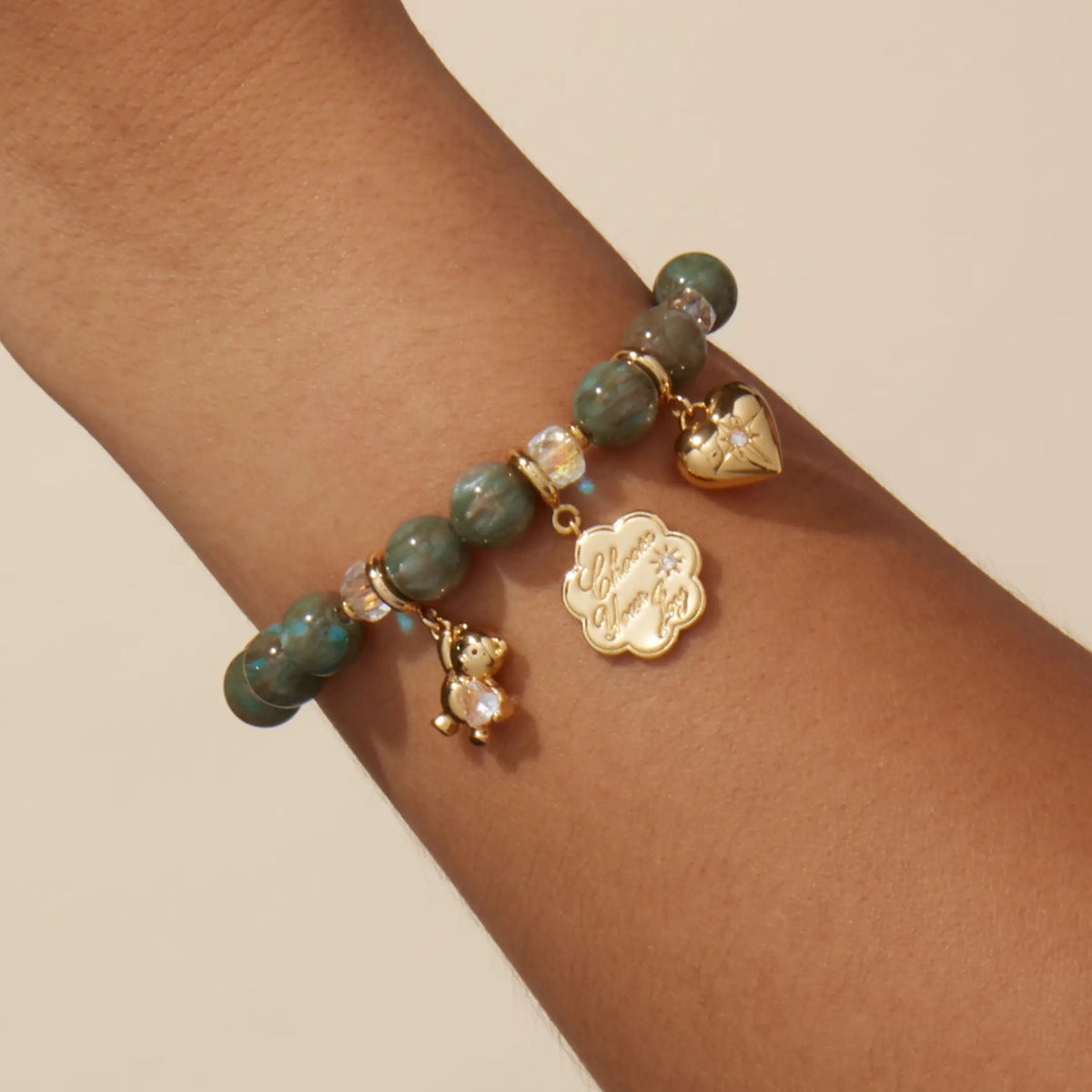 Velvet Voltage Bracelet Base worn on a wrist styled with gold teddy bear gem, Choose Your Joy coin, and cz heart charms, on-model on a tan background
