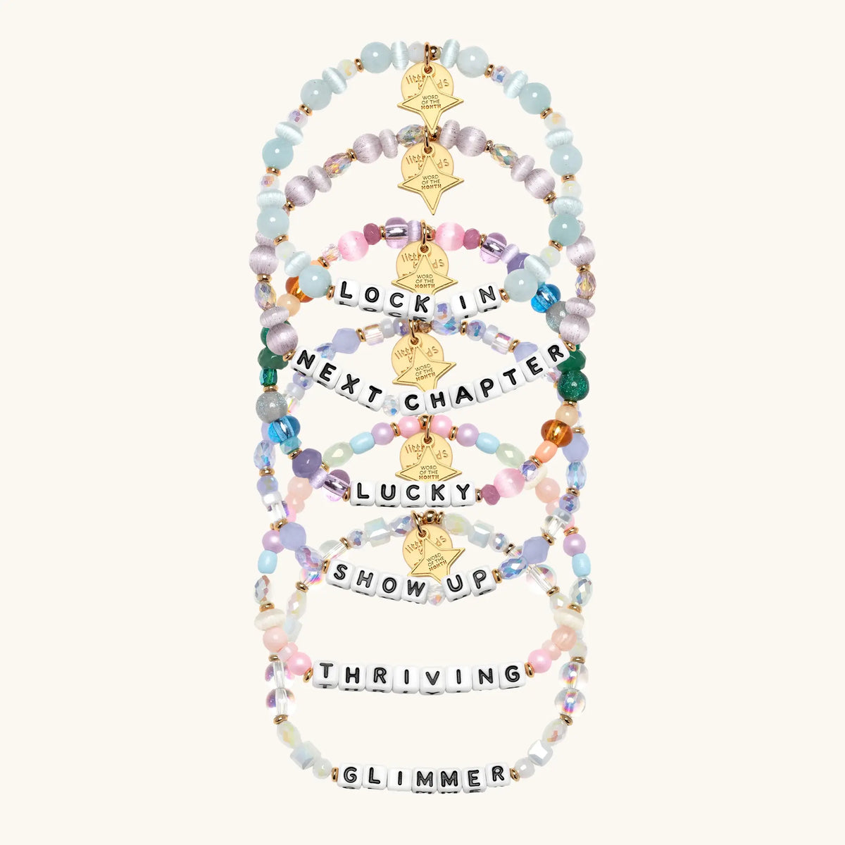 A set of six beaded stretch bracelets arranged vertically on a light cream background. The bracelets feature pastel-colored beads in shades of blue, pink, purple, and green, accented with gold star tags and letter beads spelling out motivational phrases like 'LOCK IN', 'NEXT CHAPTER', 'LUCKY', 'SHOW UP', 'THRIVING', and 'GLIMMER'.