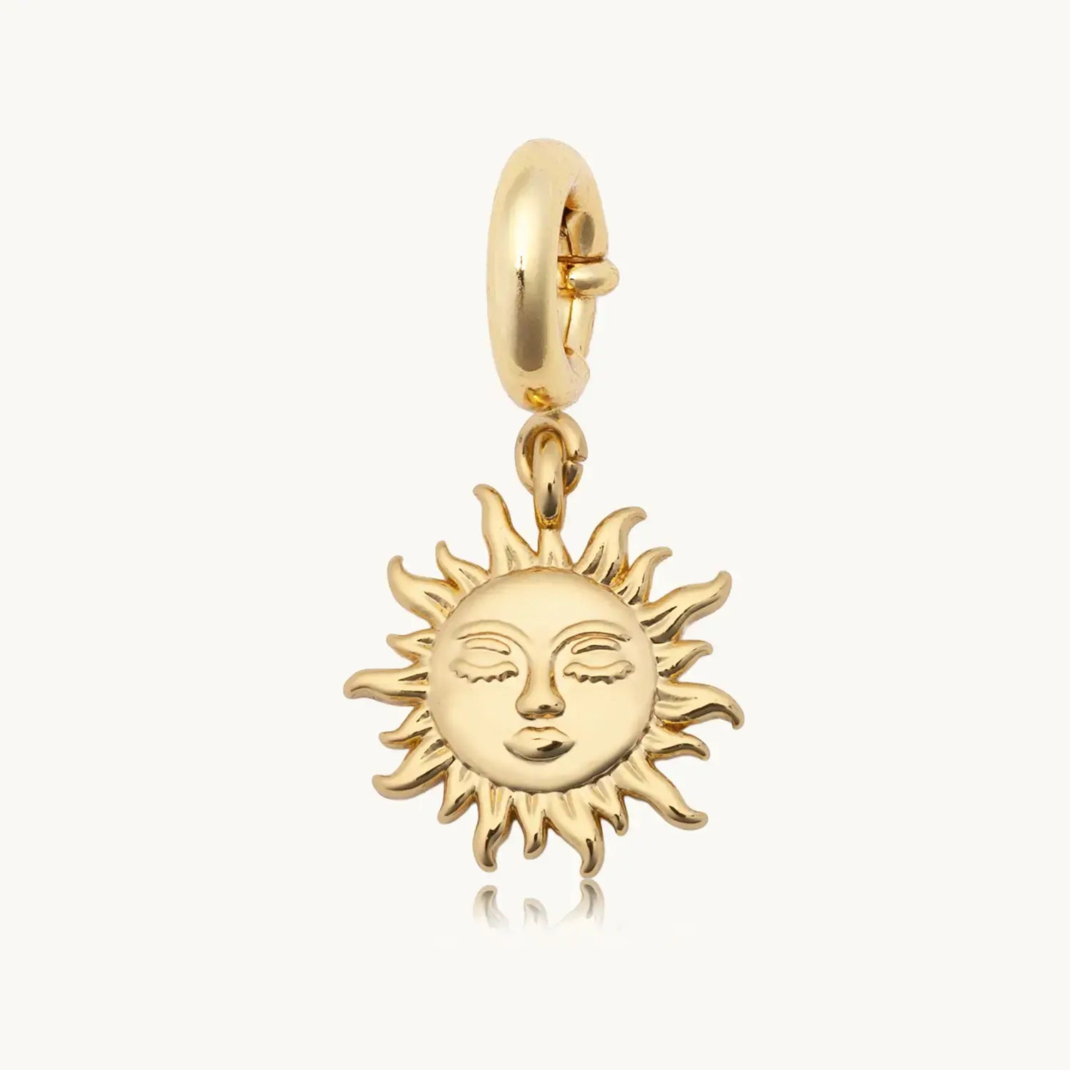 Gold sun face charm pendant with a serene, closed-eye facial expression surrounded by radiating flame-like rays, hanging from a polished gold spring-ring clasp, on a light grey background.
